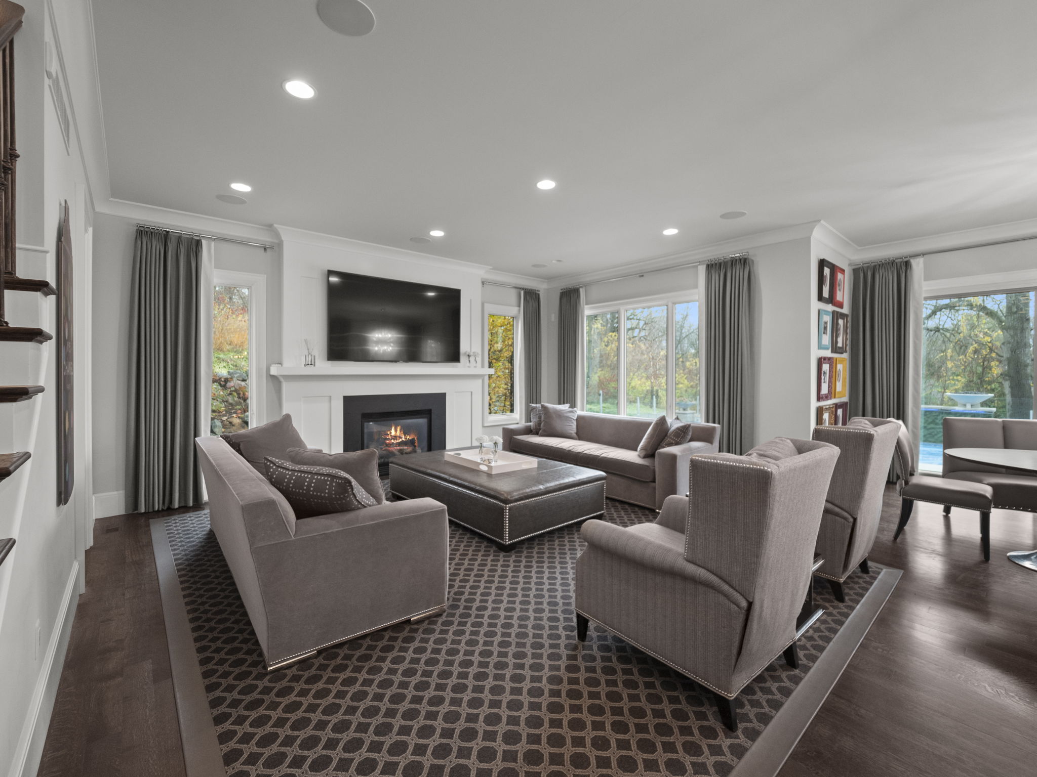 This is a well-lit living room featuring a fireplace with a large television mounted above it. The room is furnished with two sofas, two armchairs, and a large ottoman, all in a neutral color palette. The flooring is a patterned carpet, and the room has large windows with views of the outdoors, creating a comfortable and inviting atmosphere.