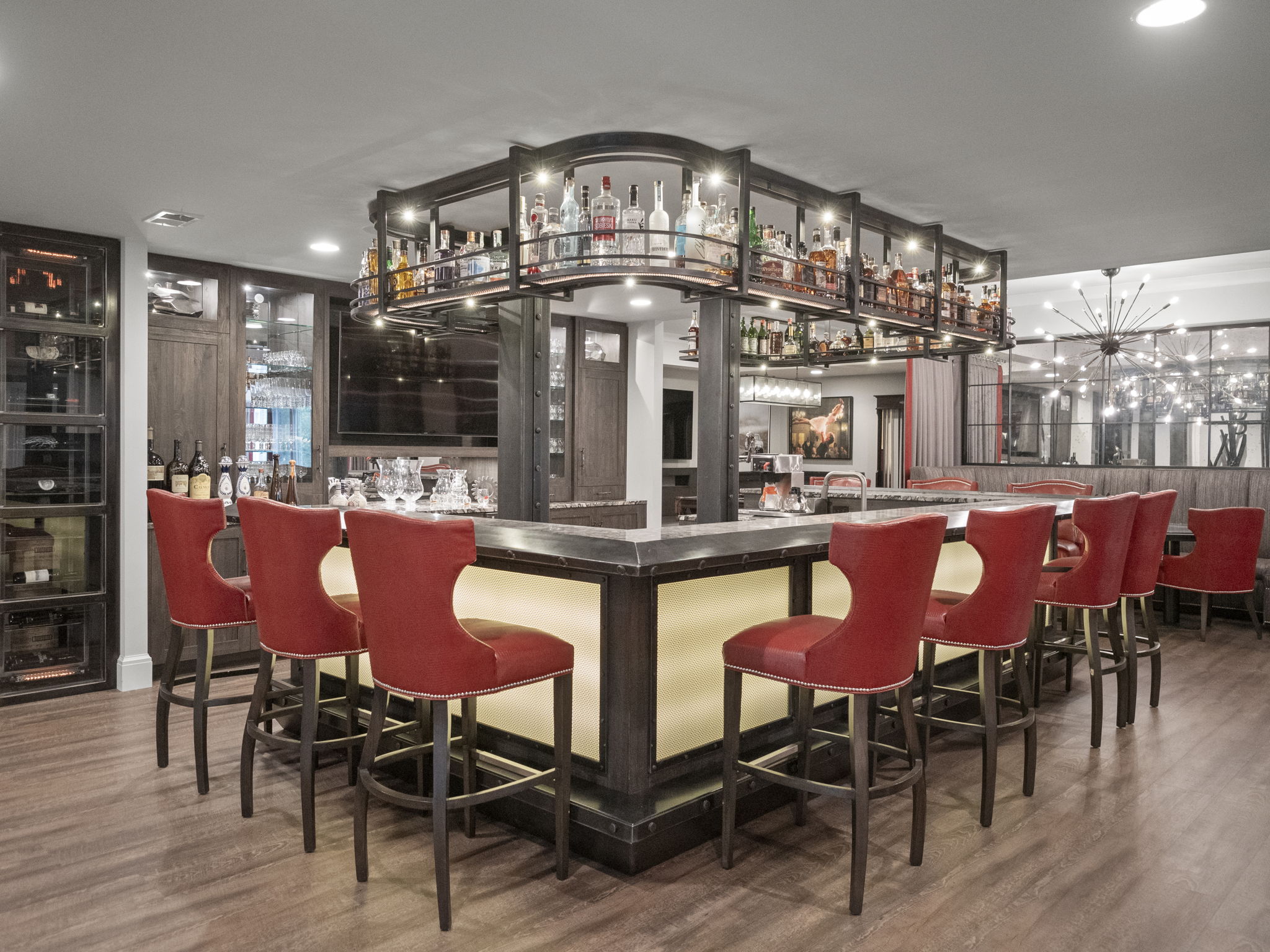 This is an interior shot of a luxurious wine cellar and bar area. The space features a well-stocked bar with overhead shelving displaying numerous bottles, surrounded by red upholstered bar stools. The room is illuminated with warm lighting, highlighting the rich wood flooring and custom cabinetry, creating an inviting and sophisticated atmosphere.