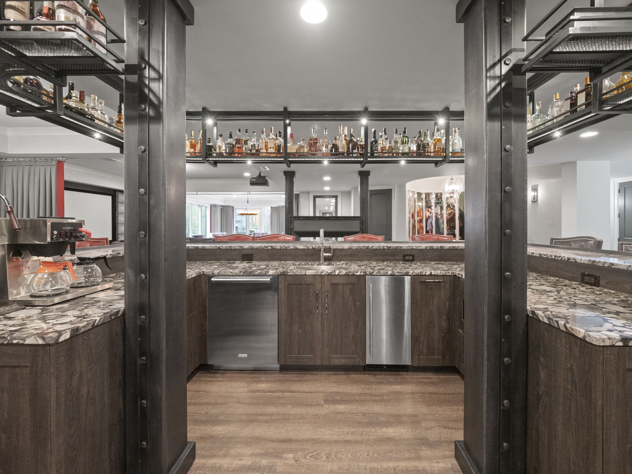 This is an interior shot of a basement bar area, featuring dark wood cabinetry, granite countertops, and stainless steel appliances. The bar is well-stocked with liquor bottles displayed on shelves above, and the space is framed by dark metal support beams, creating a sophisticated and inviting atmosphere. The flooring is a light wood laminate.