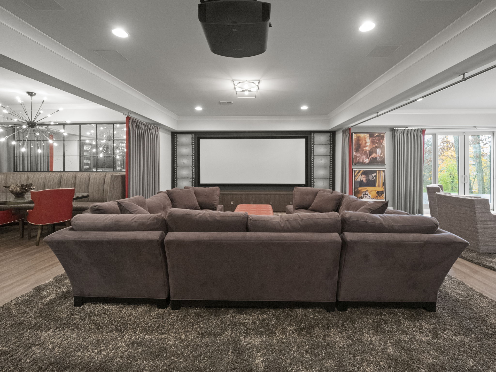 This is an interior shot of a home theater or living room, featuring a large sectional sofa in the foreground, facing a projection screen. The room is designed for entertainment, with movie posters and built-in shelving flanking the screen. The perspective is from the back of the room, looking towards the screen, creating a sense of immersion.