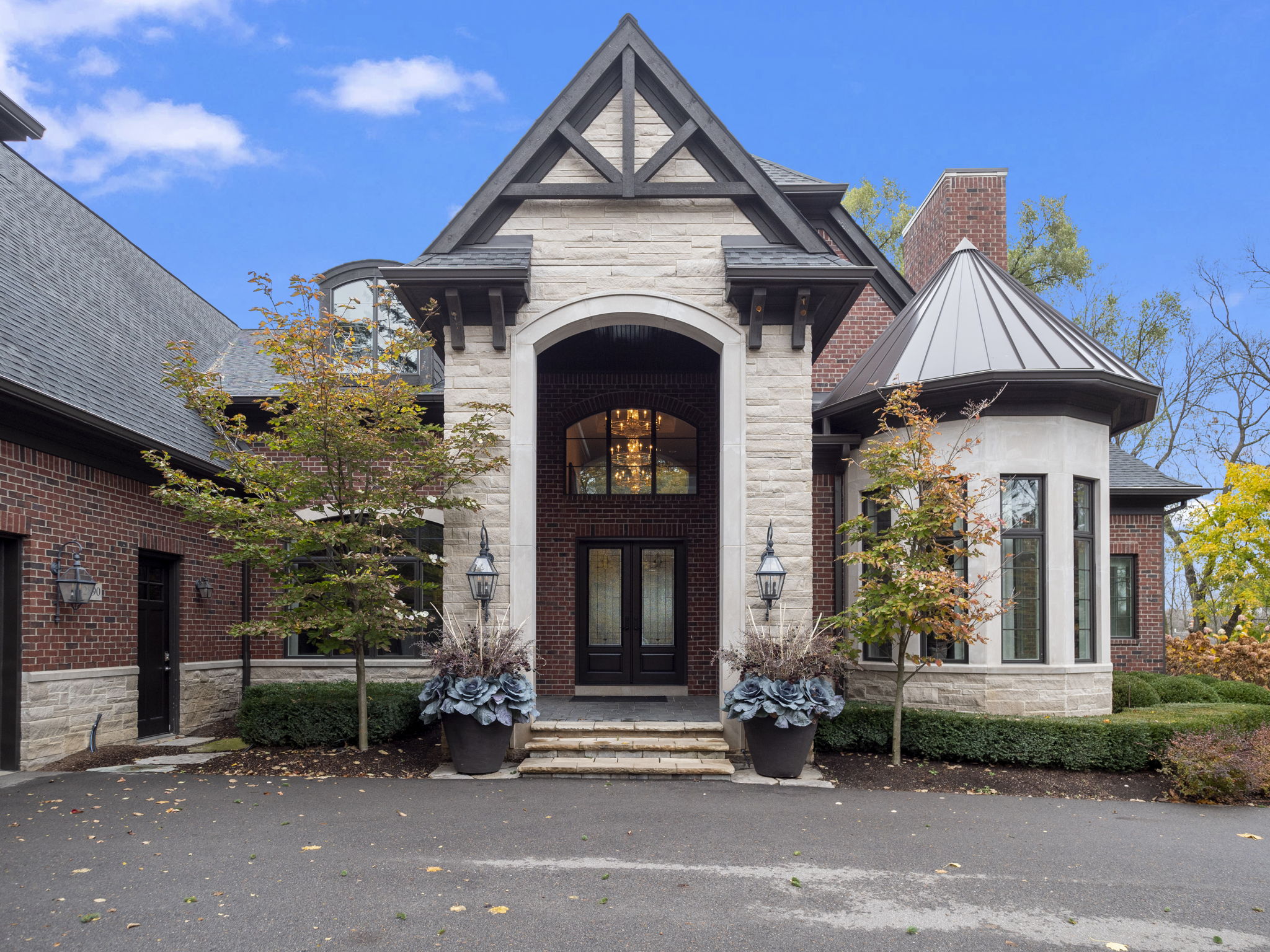 This is a stately home's entryway, featuring a stone facade, a prominent arched entrance, and a dark wood double door. The entrance is flanked by large planters and symmetrical landscaping, creating a grand and inviting first impression. The architecture combines traditional and modern elements, suggesting a luxurious and well-maintained property.