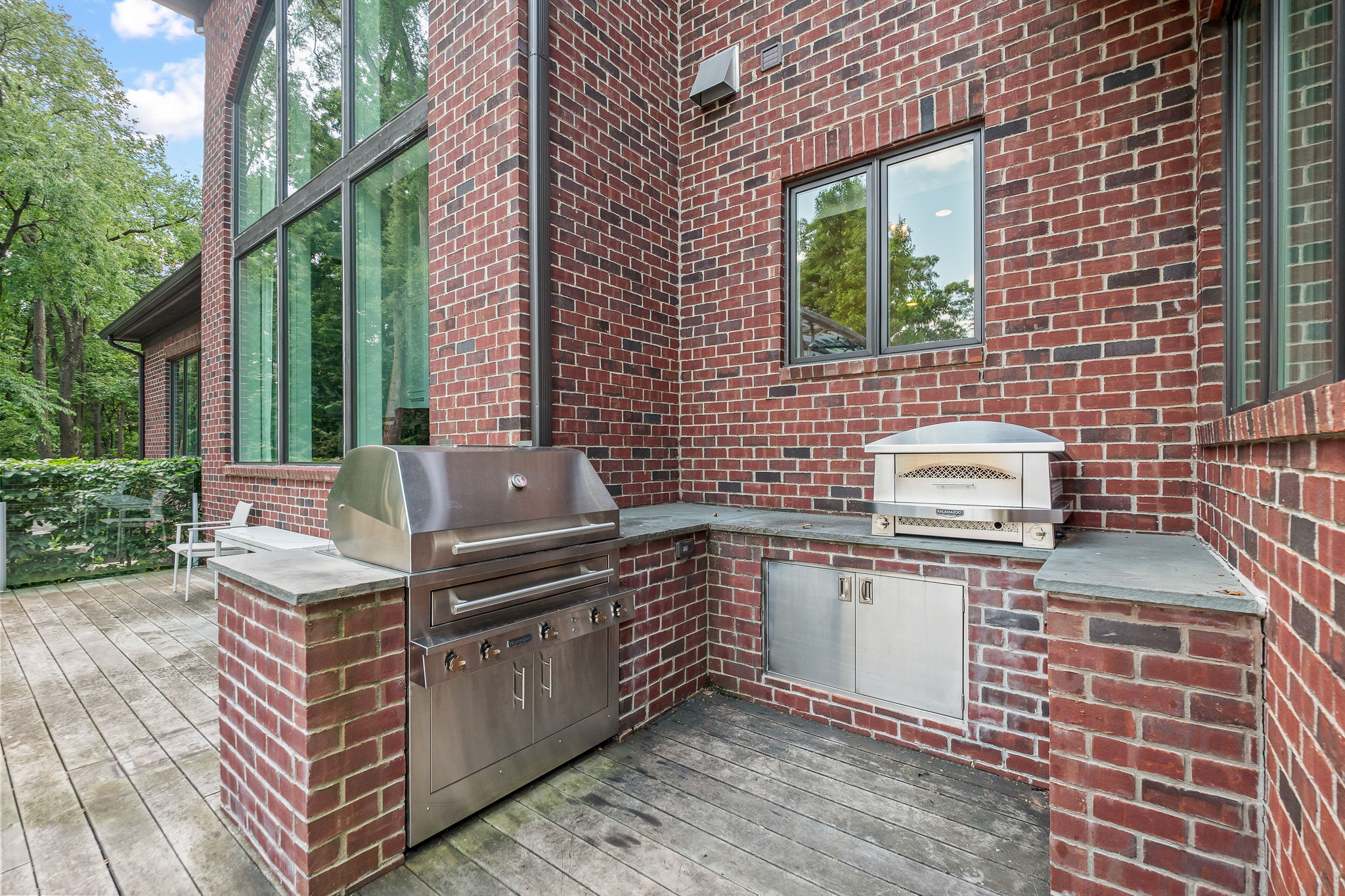This image showcases an outdoor kitchen area on a patio or deck. The space features a stainless steel grill and a pizza oven built into brick structures with stone countertops. The patio is made of wooden planks, and the area is adjacent to a brick building with large windows, creating an inviting outdoor cooking and entertaining space.