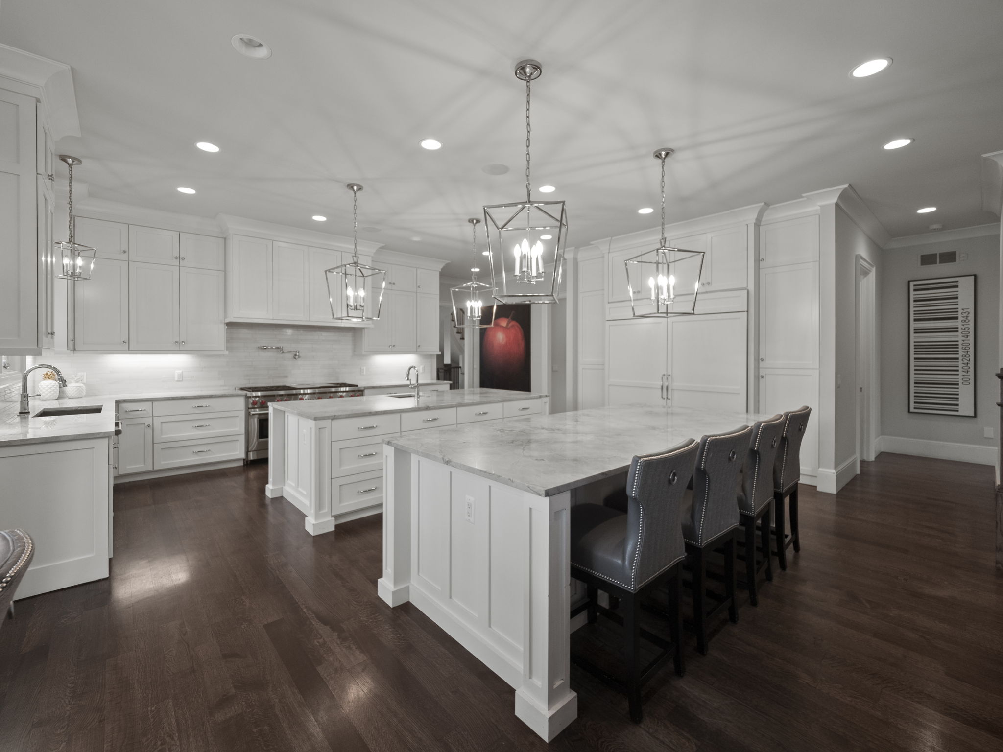 This is a bright and spacious kitchen featuring two large islands with marble countertops, white cabinetry, and stainless steel appliances. The kitchen is illuminated by pendant lighting and recessed lighting, creating a warm and inviting atmosphere. The dark hardwood floors add a touch of elegance to the space.
