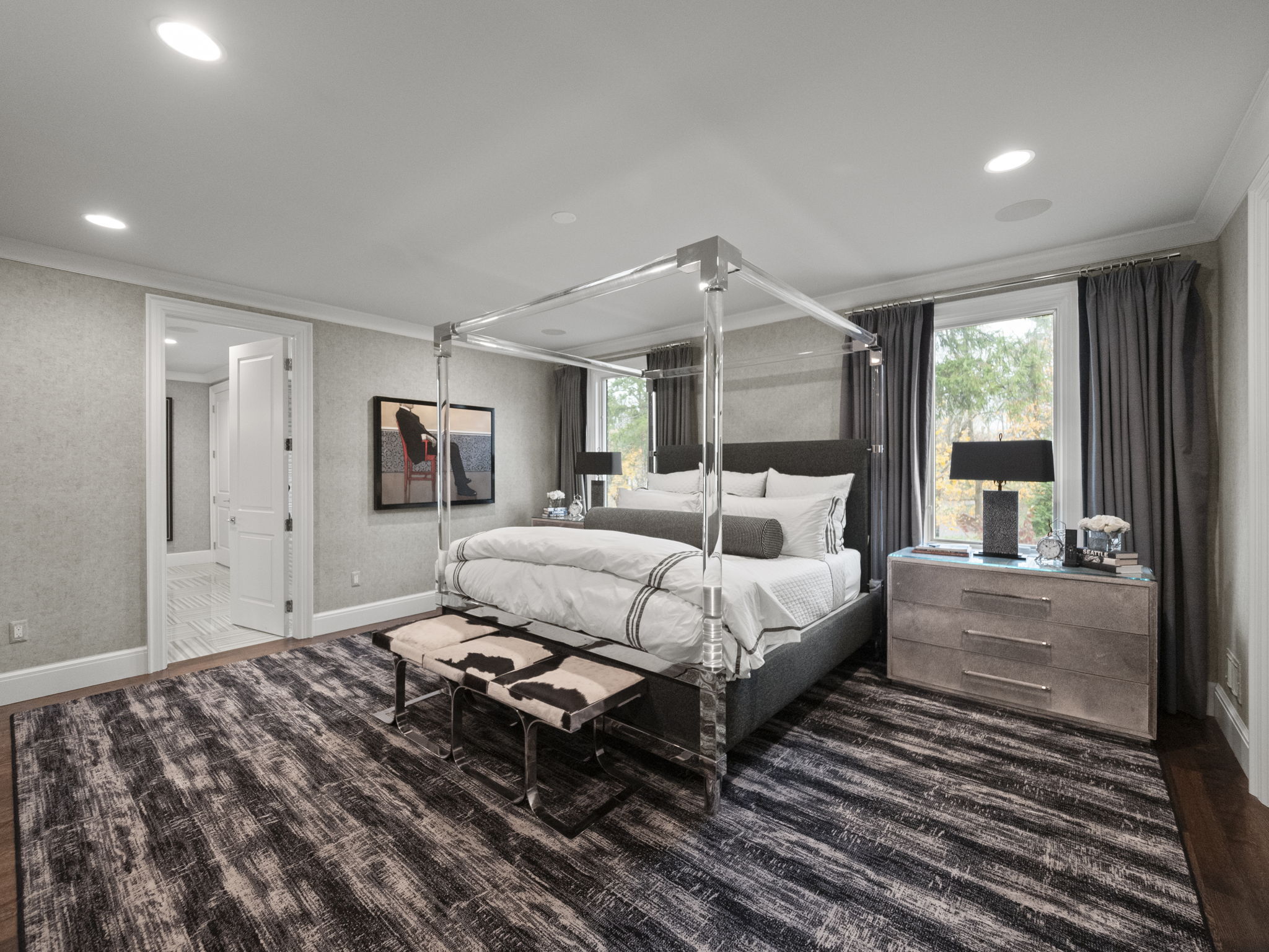 This is a primary bedroom featuring a four-poster bed with a gray upholstered frame and white bedding. The room is decorated in neutral tones with patterned wallpaper and gray curtains. A bench sits at the foot of the bed, and a nightstand with a lamp is positioned on one side. The perspective is from the doorway, showcasing the entire room.