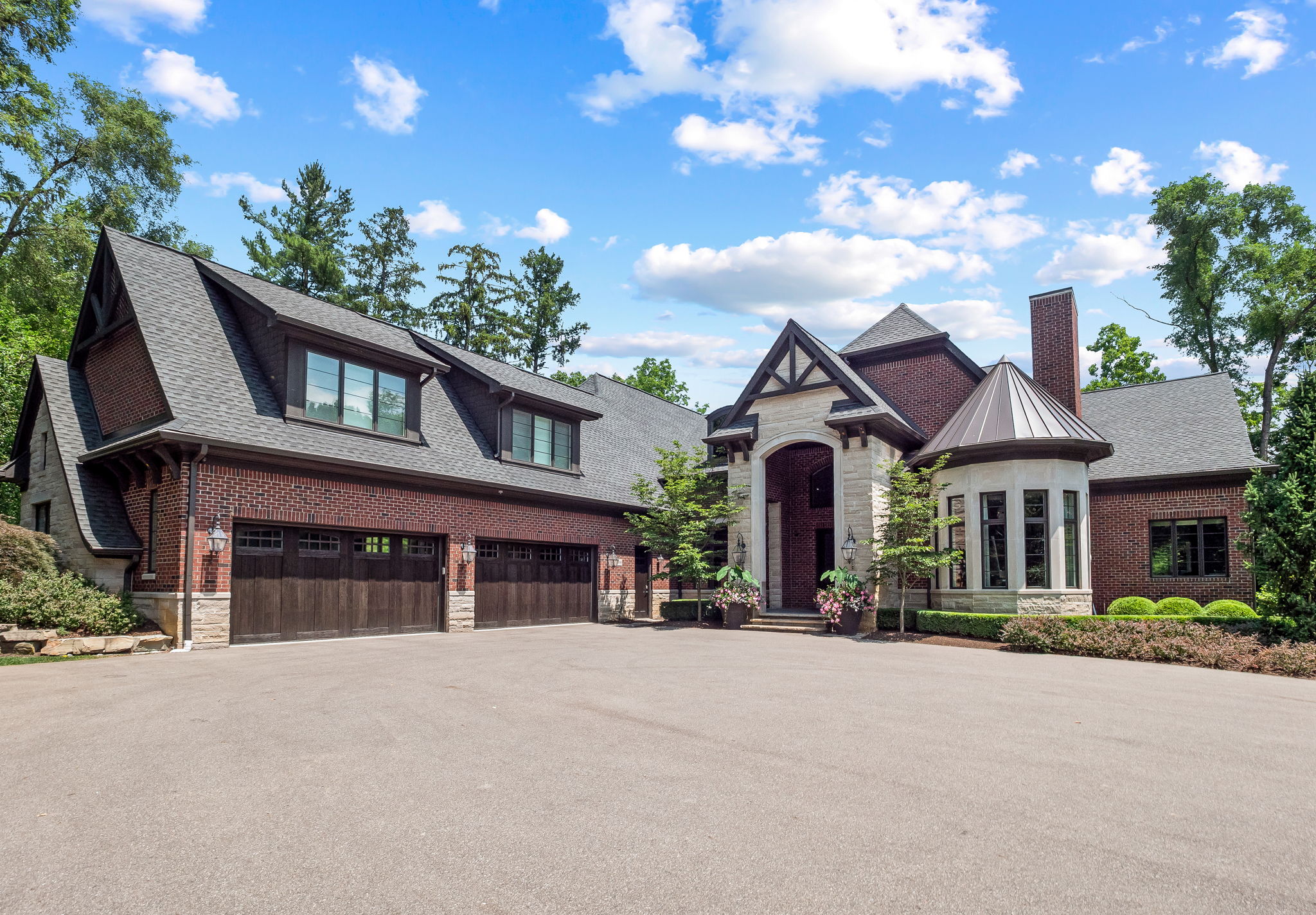 This is a front exterior view of a large, luxurious brick home with a prominent stone entryway. The house features a multi-car garage, a turret-like structure, and well-maintained landscaping. The overall impression is one of elegance and grandeur, set against a backdrop of mature trees and a blue sky with scattered clouds.