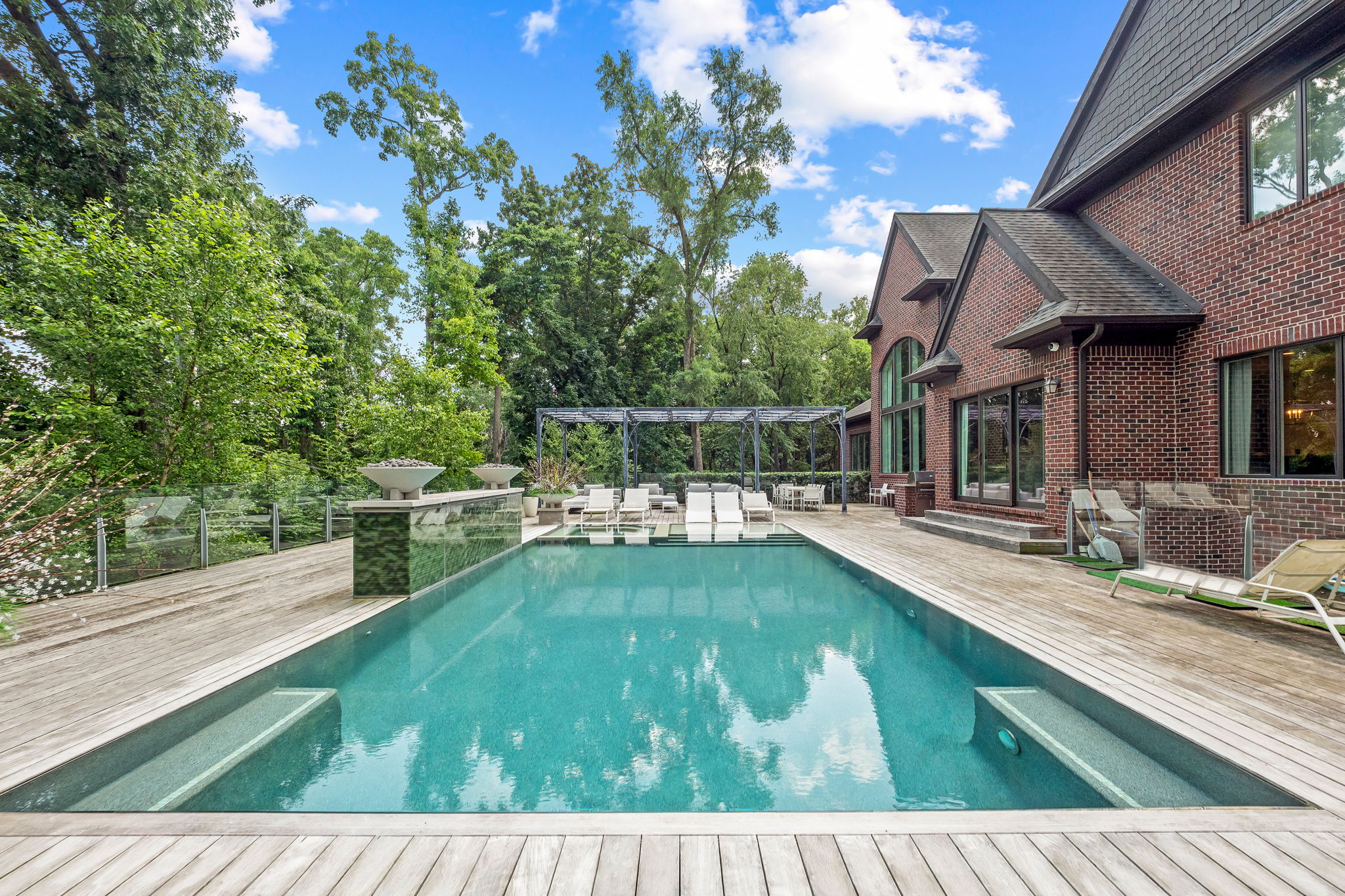 This image showcases a luxurious backyard pool area, featuring a rectangular pool with built-in steps and a wooden deck. Lounge chairs are arranged under a pergola, creating a relaxing space for sunbathing and entertaining. The backdrop includes lush greenery and a brick house, adding to the property's appeal.