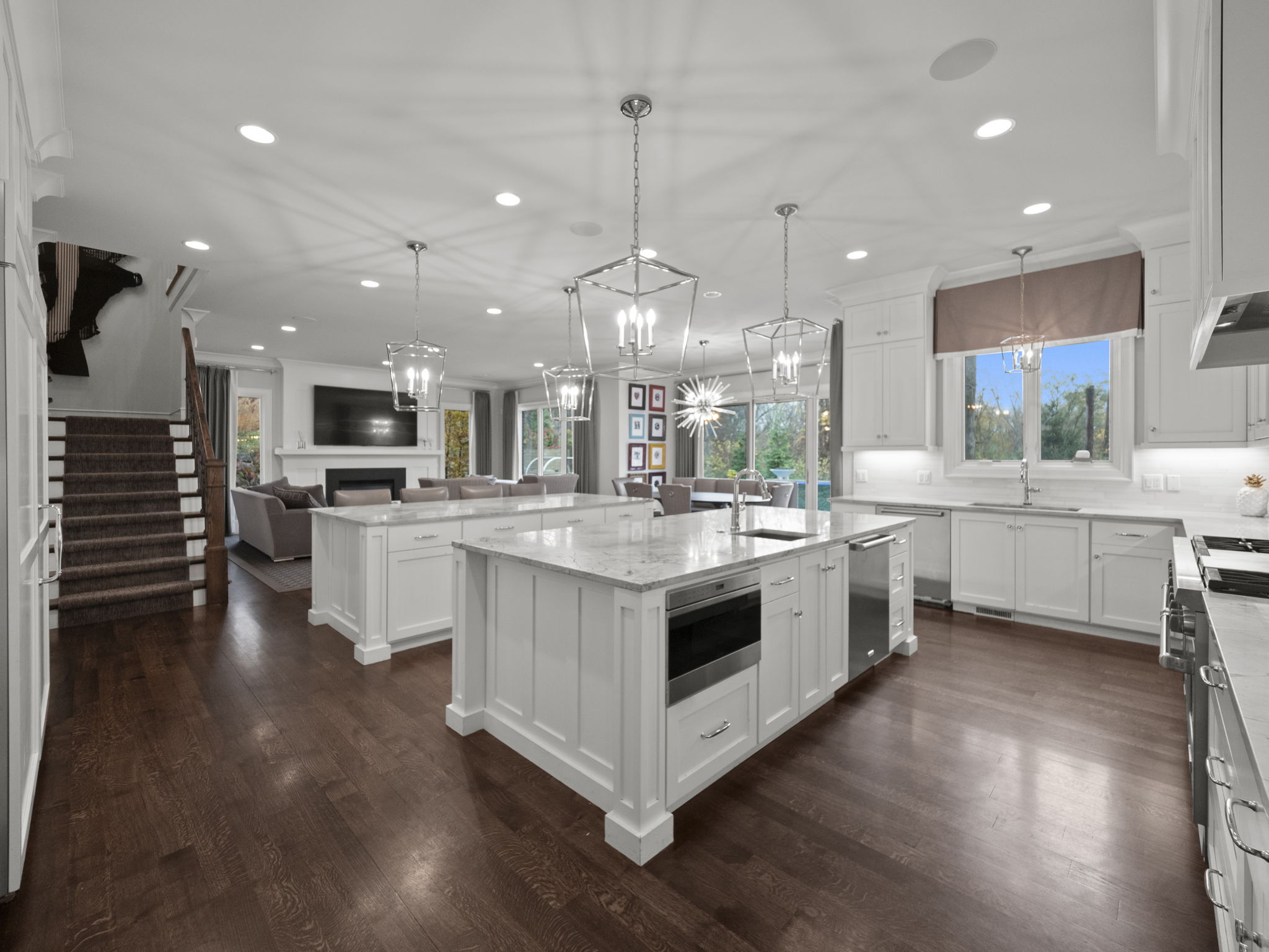 This is a spacious, open-concept kitchen featuring two large islands with white cabinetry and marble countertops. The kitchen is well-lit with recessed lighting and pendant lights, and it seamlessly flows into the living and dining areas. Dark hardwood floors add warmth and contrast to the bright, modern space, creating an inviting atmosphere.