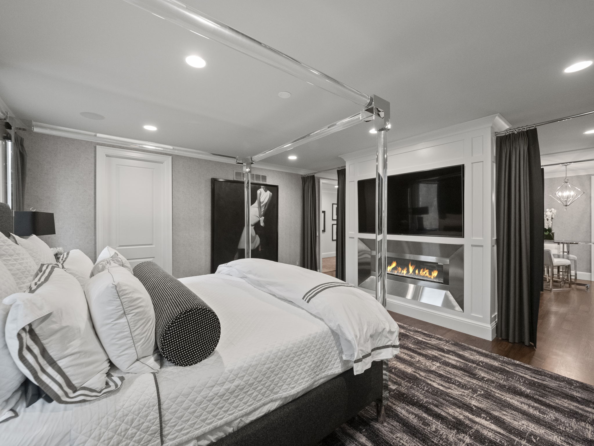 This is a luxurious primary bedroom featuring a modern four-poster bed with plush pillows and a quilted comforter. A sleek fireplace and television are integrated into a wall unit, adding a touch of elegance and entertainment. The room is well-lit with recessed lighting and features a neutral color palette, creating a serene and inviting atmosphere.