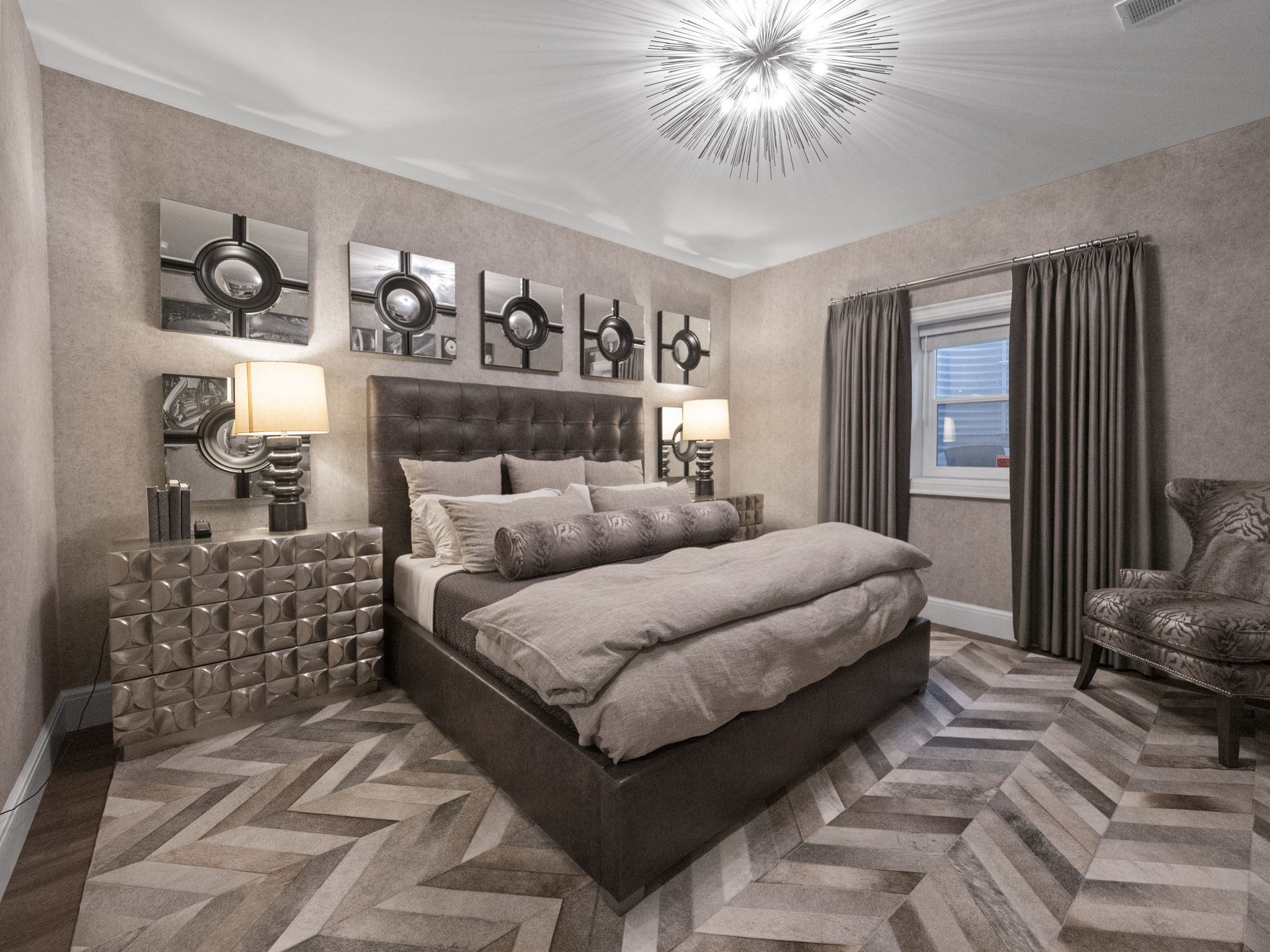 This is a luxurious primary bedroom featuring a large bed with a tufted headboard and neutral-toned bedding. The room is decorated with modern square mirrors above the bed and a unique dresser with a geometric design. A striking chandelier illuminates the space, and the herringbone-patterned floor adds a touch of elegance.