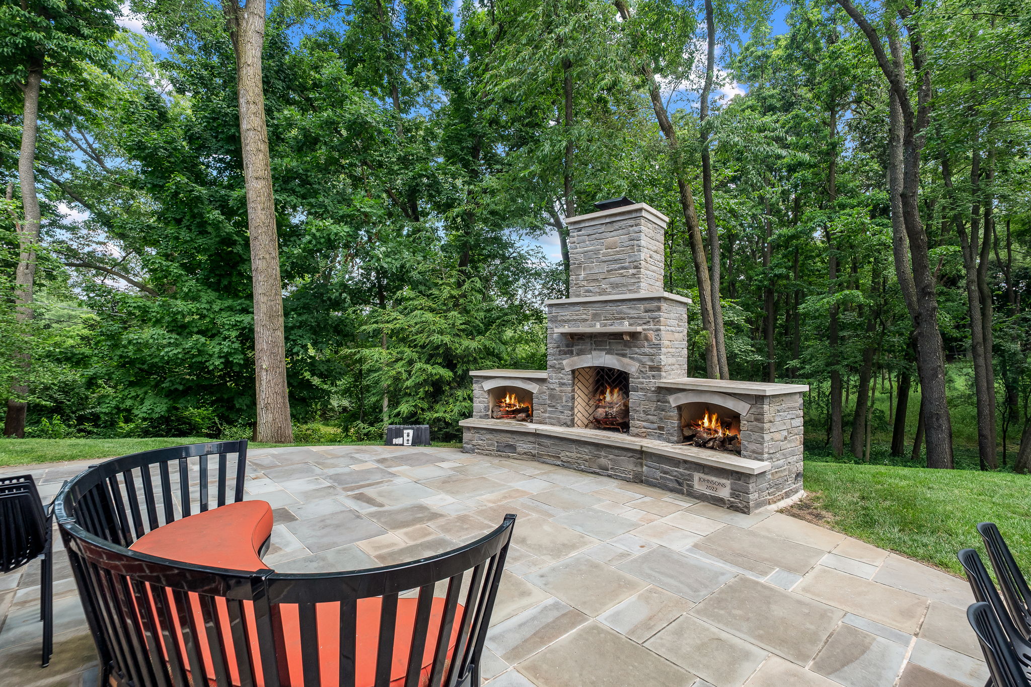 This image showcases a beautifully designed outdoor patio area, featuring a stone fireplace with three openings and a paved stone patio. The patio is furnished with curved black chairs with orange cushions, creating a cozy and inviting atmosphere. Lush greenery and trees surround the space, providing privacy and a serene backdrop.
