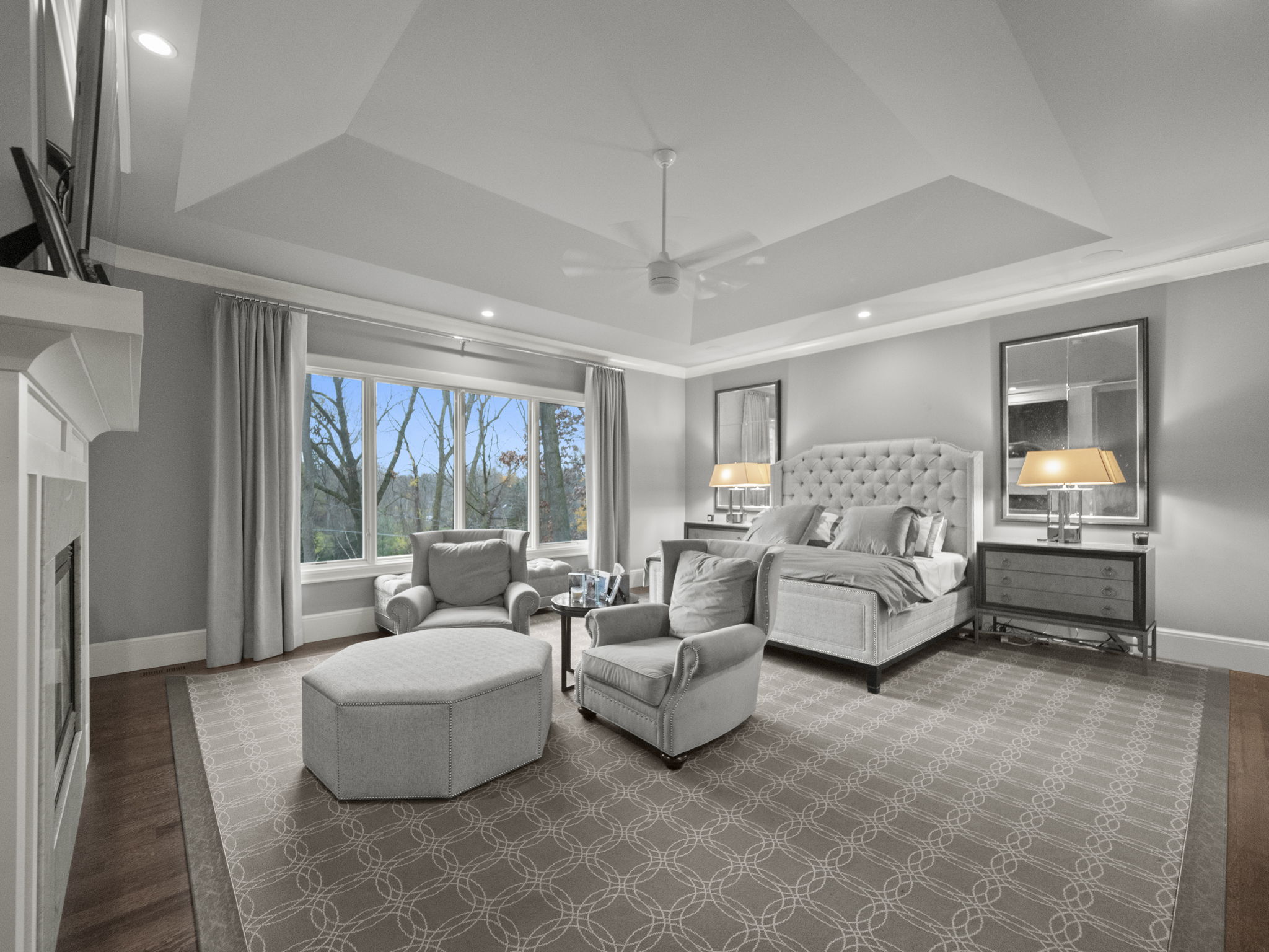 This is a spacious primary bedroom featuring a tufted headboard bed, two armchairs, and an ottoman, creating a comfortable seating area. The room is decorated in neutral tones with a patterned rug and ample natural light from a large window. The tray ceiling and fireplace add architectural interest, enhancing the luxurious feel of the space.