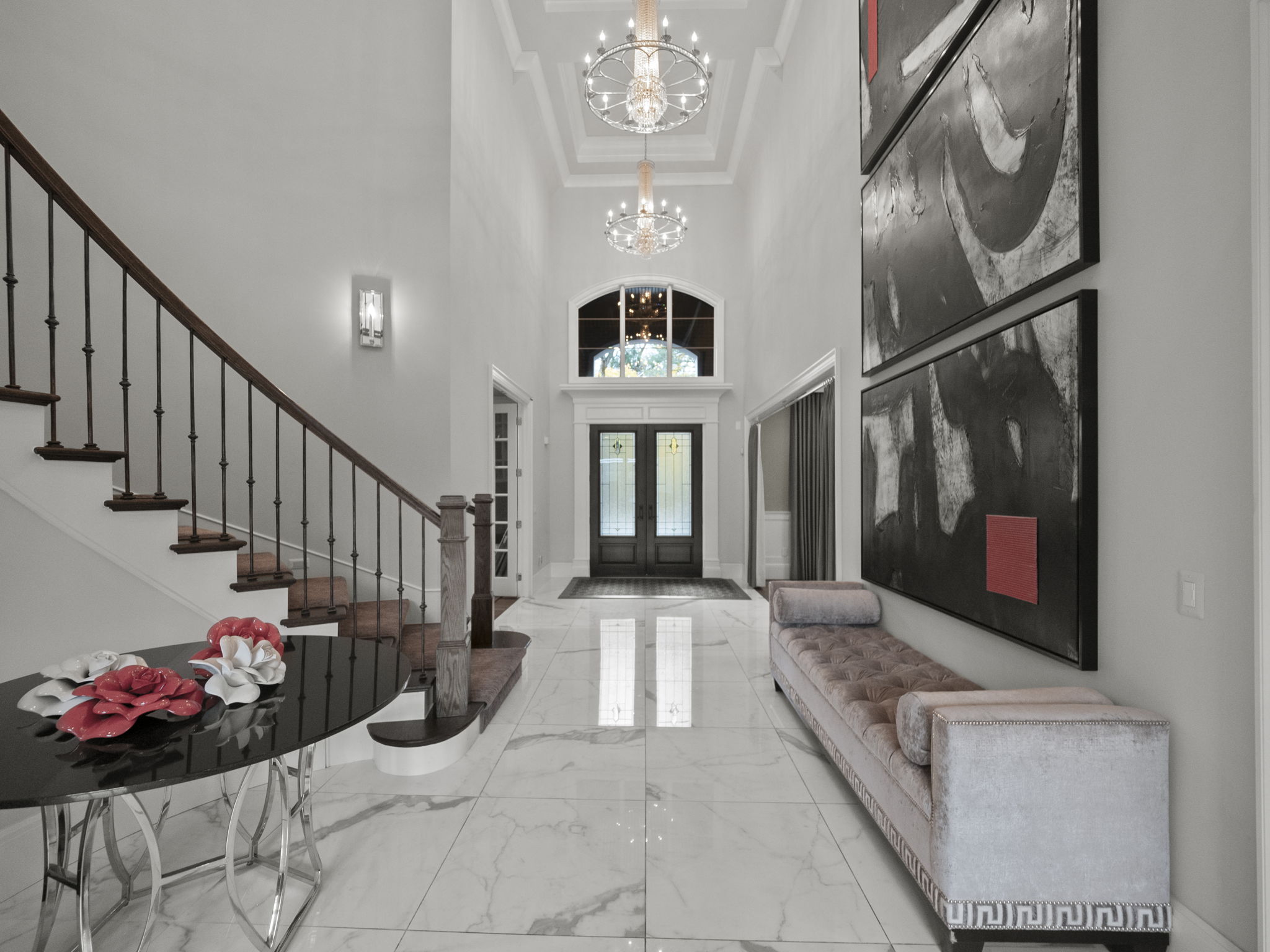 This is an interior shot of a grand foyer featuring a staircase, marble flooring, and elegant lighting fixtures. A long bench sits against the wall, complemented by modern artwork. The space is bright and airy, creating a luxurious and welcoming atmosphere.