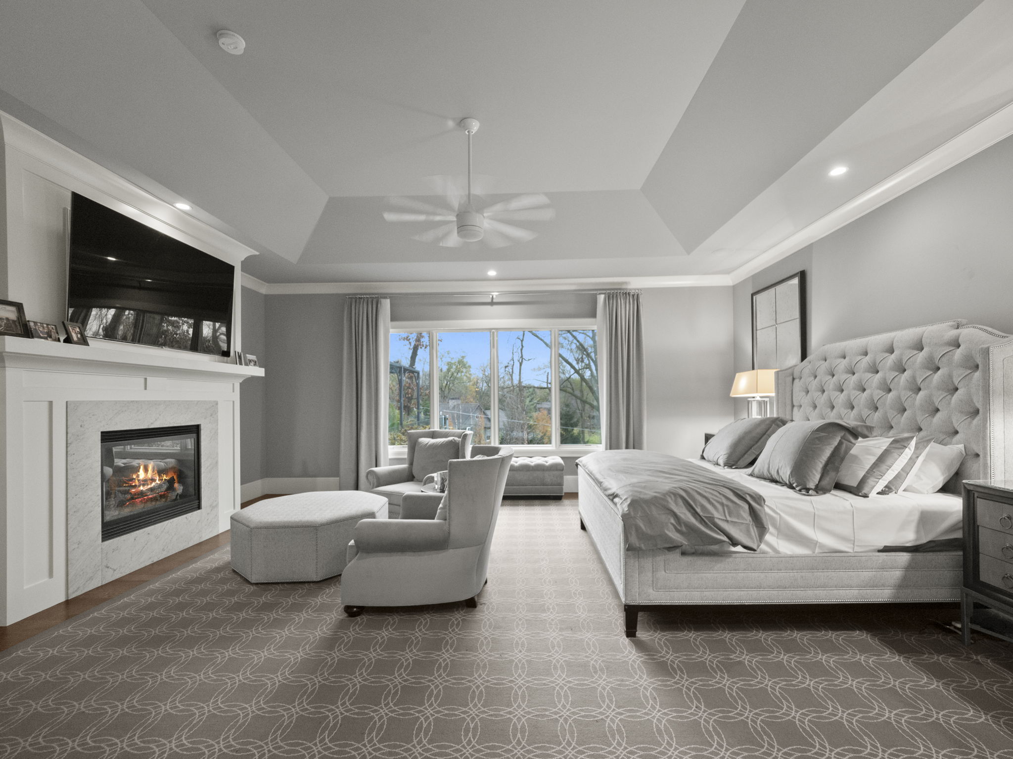 This is a spacious primary bedroom featuring a tray ceiling with a ceiling fan, a fireplace with a television above, and a seating area by a large window. The room is decorated in neutral tones, with a large upholstered bed and matching furniture. The overall impression is luxurious and comfortable.