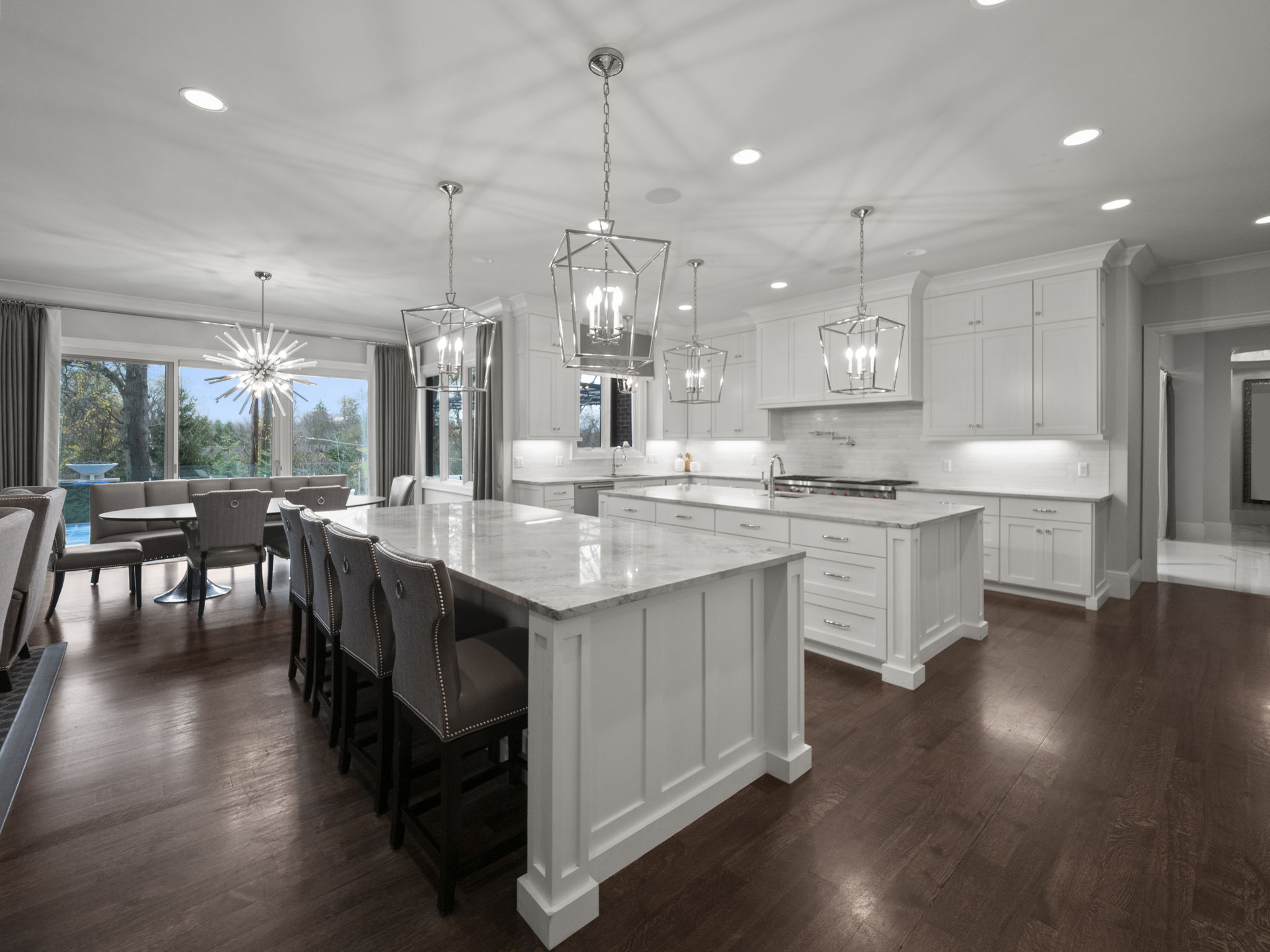 This is a bright and spacious kitchen featuring two large islands with white marble countertops and white cabinetry. The kitchen is illuminated by pendant lighting and recessed lighting, creating a warm and inviting atmosphere. The dark hardwood floors add a touch of elegance and contrast to the overall design.