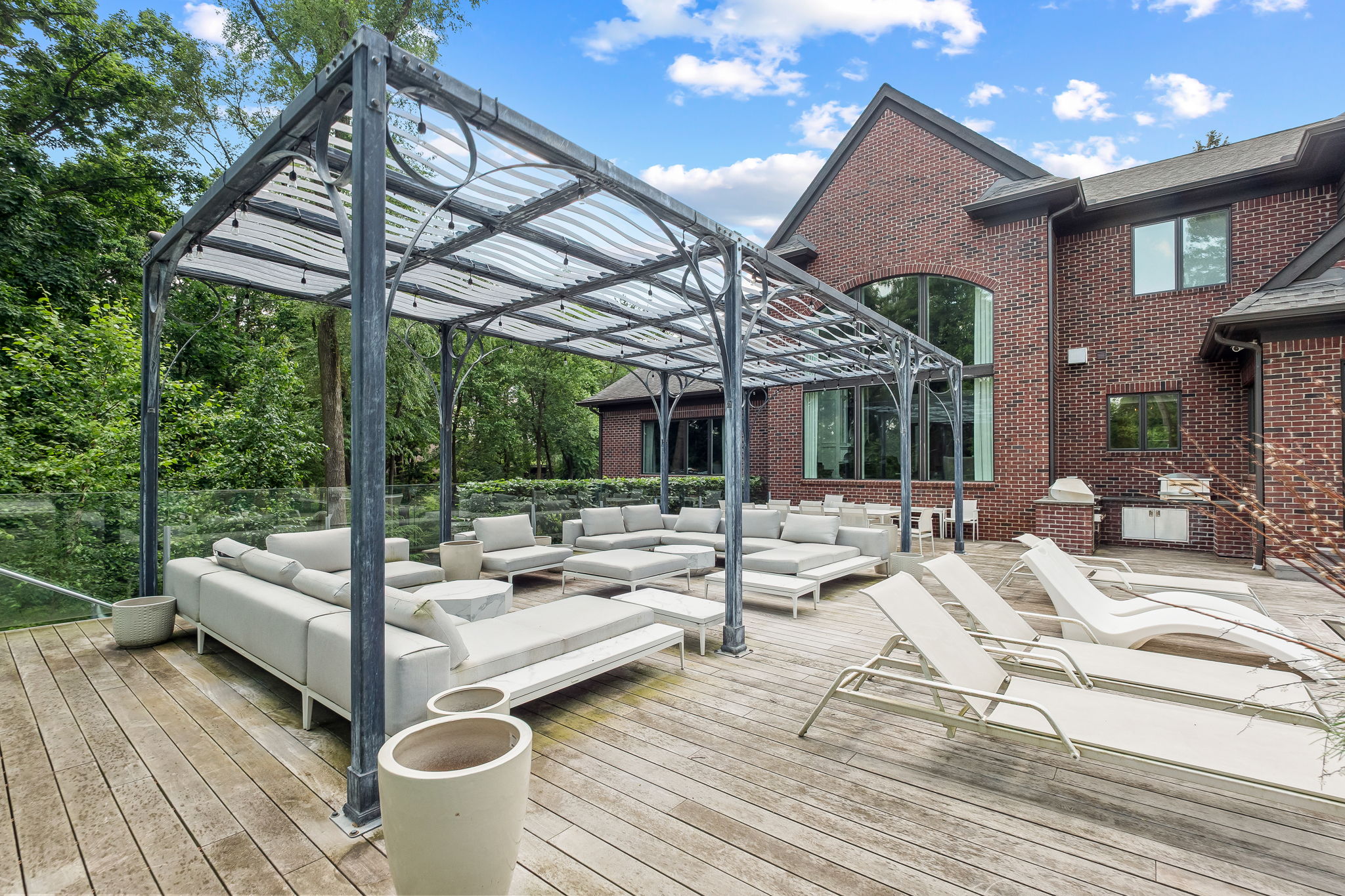 This image showcases a luxurious outdoor living space featuring a spacious wooden deck with modern, light-colored sectional seating and lounge chairs. A stylish pergola provides partial shade, while the backdrop includes a well-maintained brick house and lush greenery. The scene exudes relaxation and upscale outdoor entertainment.