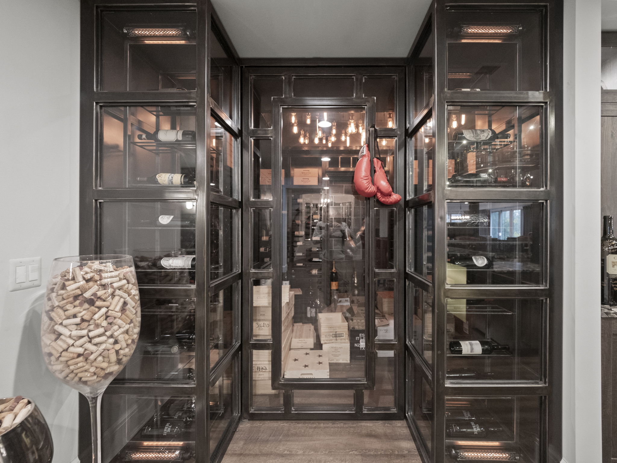 This image showcases a sophisticated wine cellar, featuring a custom-built, dark metal-framed glass enclosure. Inside, wine bottles are neatly arranged on shelves, and wooden cases are stacked on the floor. A pair of red boxing gloves hangs inside the cellar, adding a unique touch. The lighting is warm and inviting, highlighting the collection and creating an elegant atmosphere.