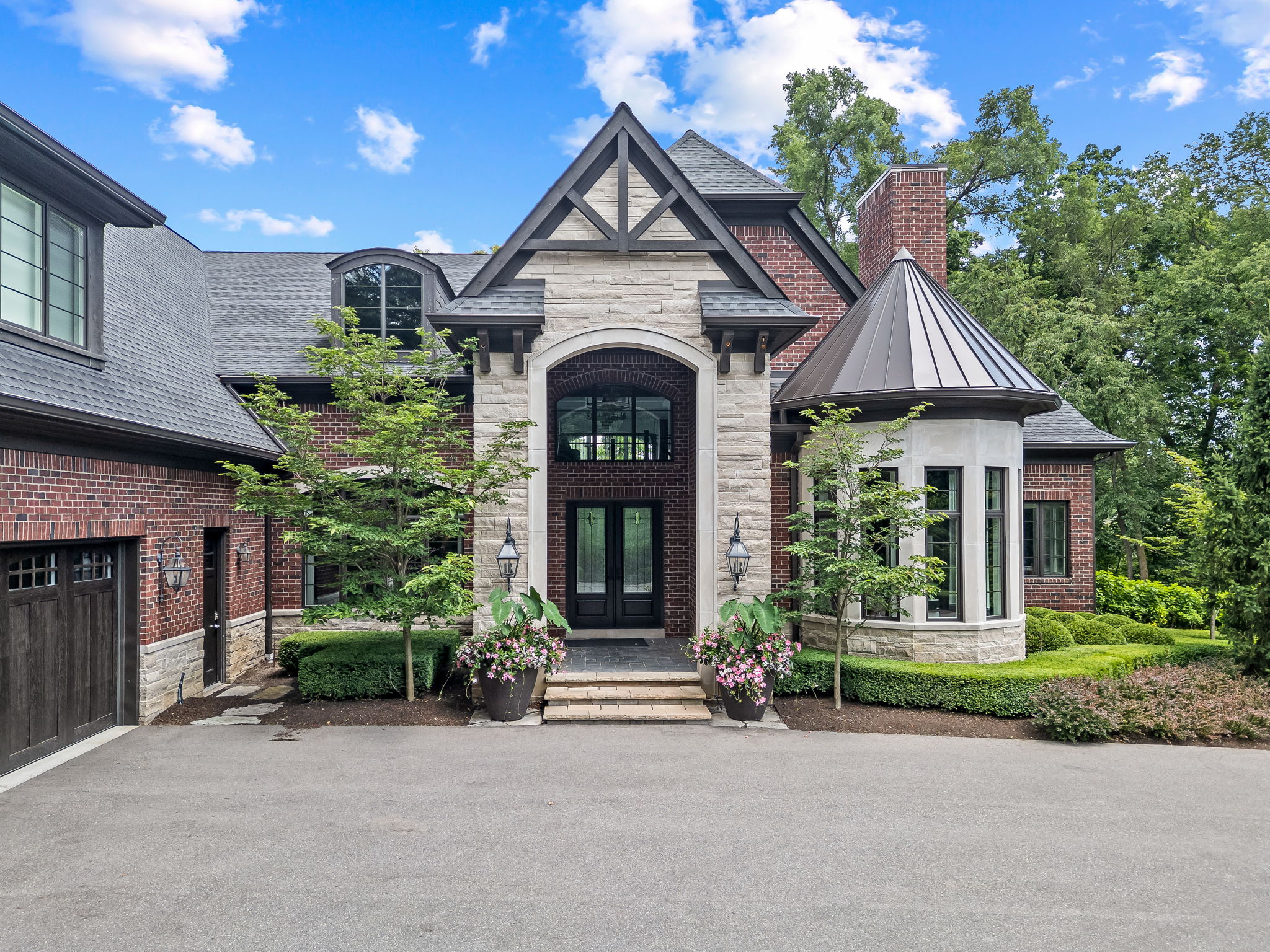 This is a front exterior view of a luxurious brick and stone home. The house features a prominent entryway with a stone archway, a dark-colored roof, and well-manicured landscaping. The overall impression is one of elegance and high-end living.