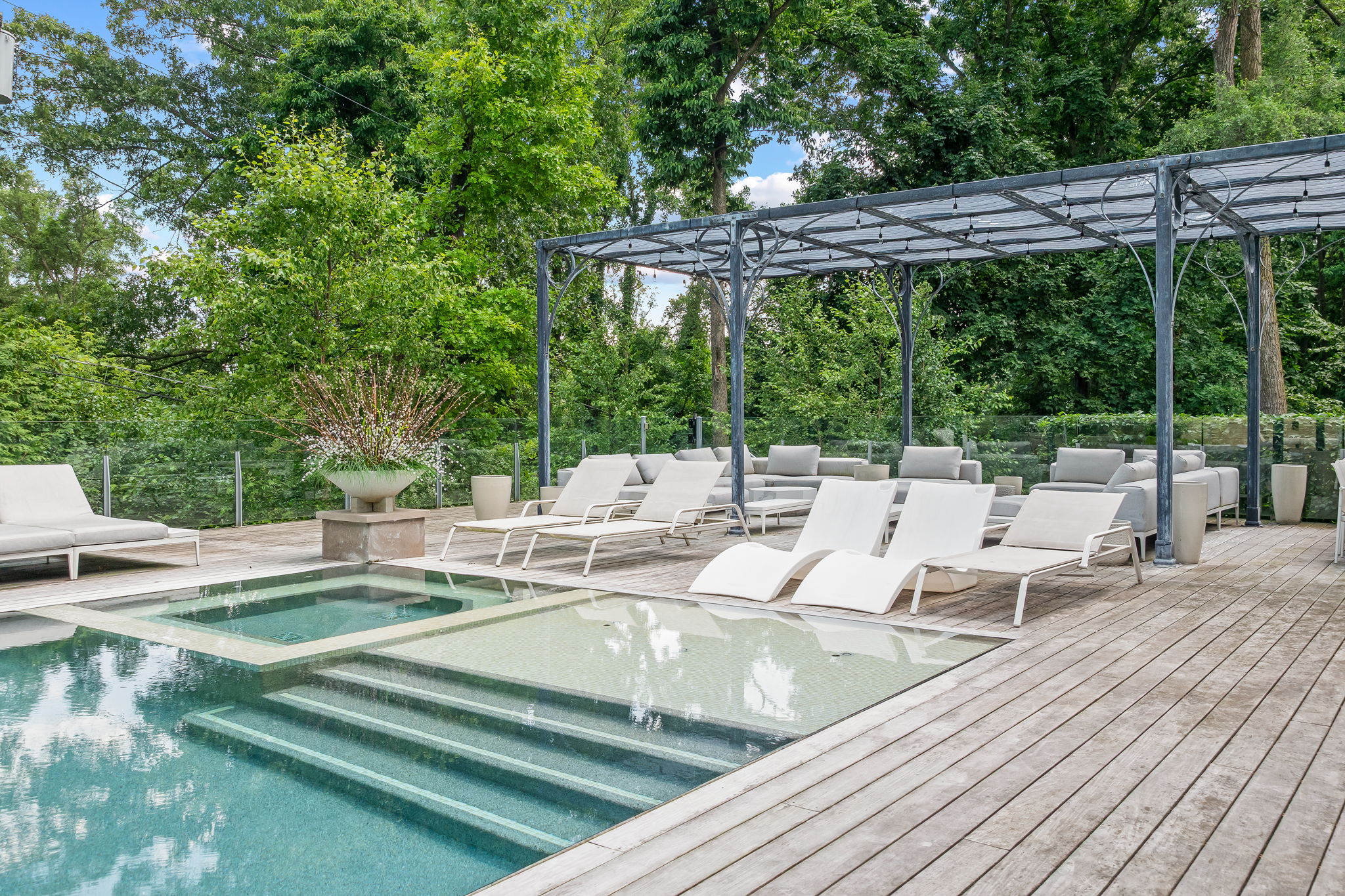 This image showcases a luxurious outdoor pool and spa area, featuring a modern design with clean lines and a wooden deck. The pool has integrated steps and a shallow area, while lounge chairs and outdoor seating provide ample space for relaxation. A pergola offers shade, and lush greenery surrounds the area, creating a private and serene atmosphere.