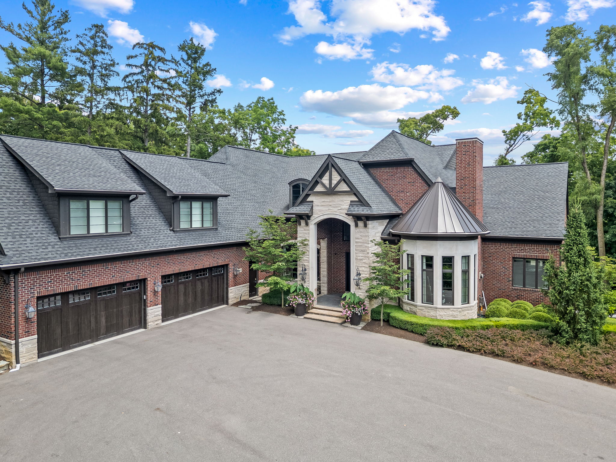 This is an eye-level, front-facing shot of a grand brick home with a dark gray roof and a spacious driveway. The house features a stone entryway, a turret-style window, and a two-car garage. The landscaping is meticulously maintained, adding to the property's curb appeal and luxurious feel.