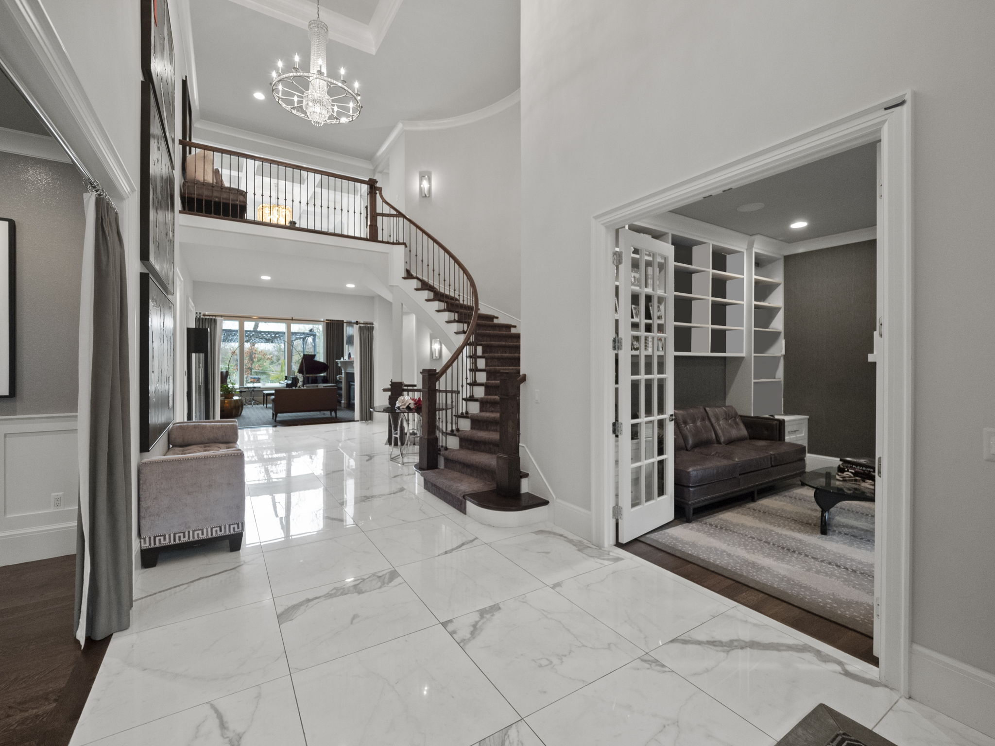 This grand foyer showcases a stunning curved staircase with dark wood treads and a wrought iron railing, illuminated by a sparkling chandelier. The space features white marble flooring, high ceilings, and an open layout that leads into adjacent living areas. A glimpse into a cozy living room with a brown leather sofa and built-in shelving adds to the home's appeal.