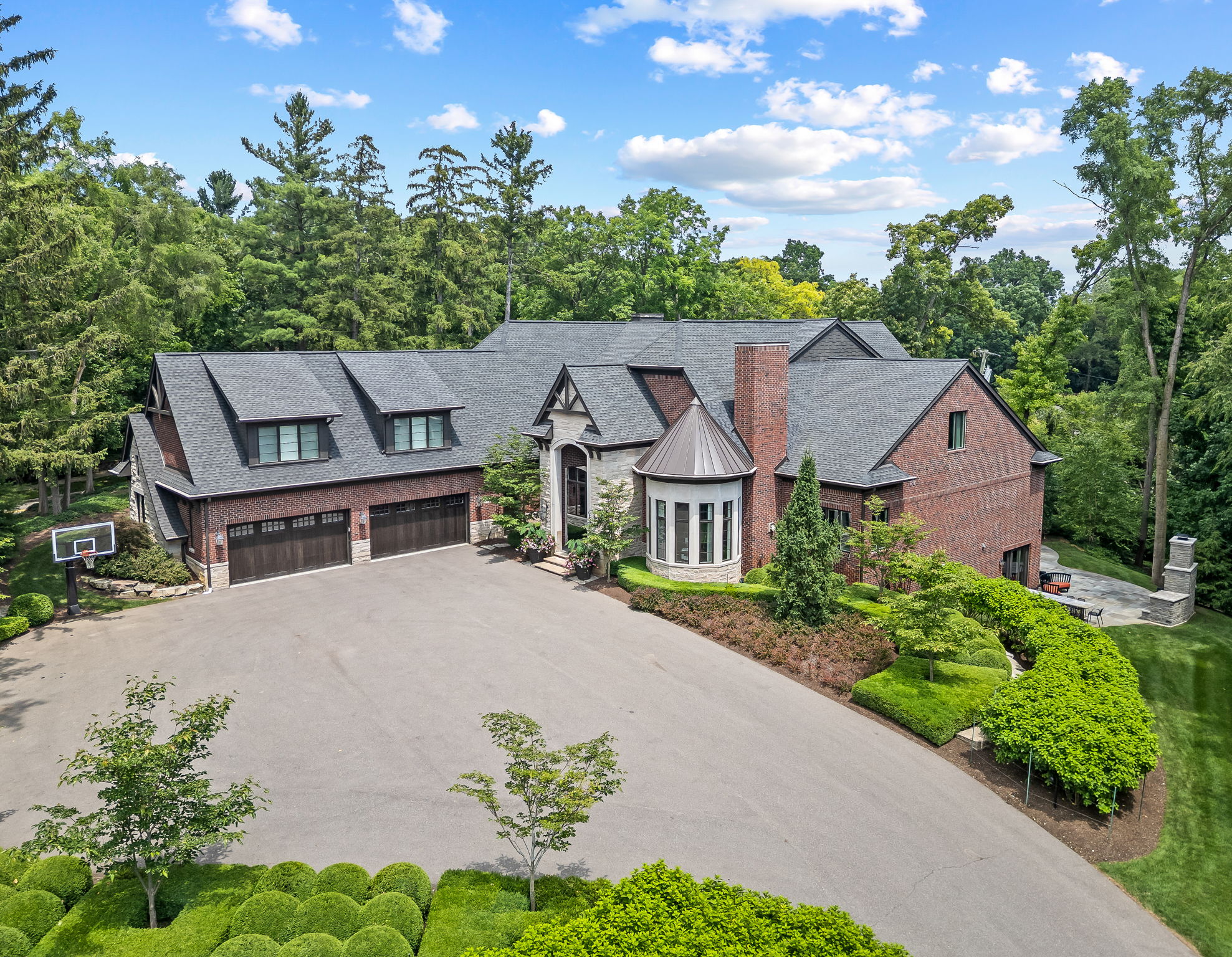 This aerial view showcases a grand brick home with a dark gray roof, multiple dormers, and a circular turret-like feature. The property boasts a large paved driveway, a basketball hoop, and meticulously landscaped grounds with mature trees and manicured shrubbery. The overall impression is one of luxury and privacy.