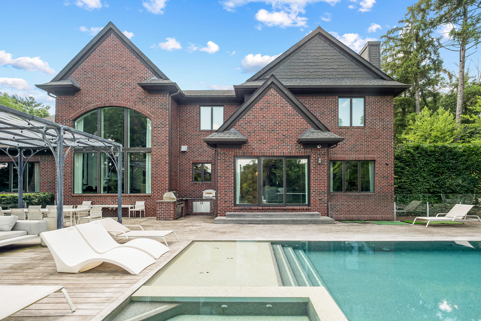 This image showcases the rear exterior of a brick house, highlighting a luxurious pool and spa area. The wooden deck features modern lounge chairs and an outdoor dining set under a pergola. The well-maintained lawn and mature trees create a private and serene backyard oasis.
