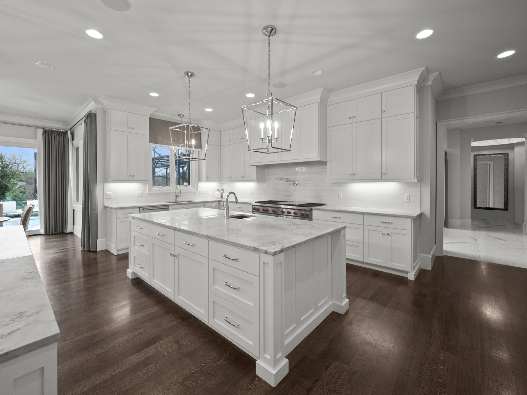 This is a bright and spacious kitchen featuring white cabinetry, a large central island with a marble countertop, and stainless steel appliances. The kitchen is illuminated by recessed lighting and pendant lights above the island, creating a warm and inviting atmosphere. Dark hardwood floors add a touch of elegance and contrast to the light color palette.