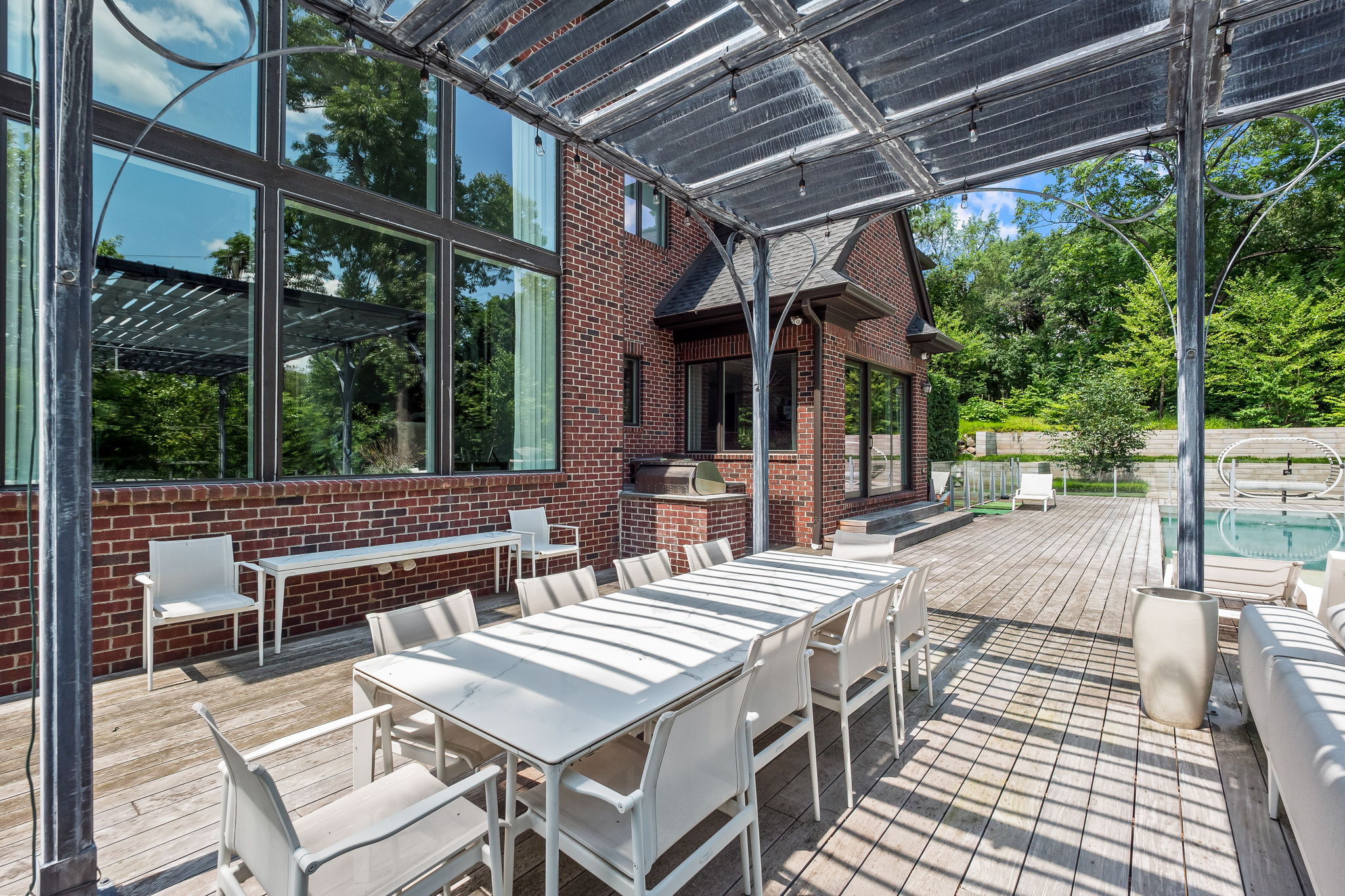 This image showcases a beautifully designed outdoor patio and deck area. A large white dining table with matching chairs sits under a modern pergola, providing shade and style. The deck extends to a pool area, creating a seamless transition between dining and relaxation spaces, perfect for entertaining.