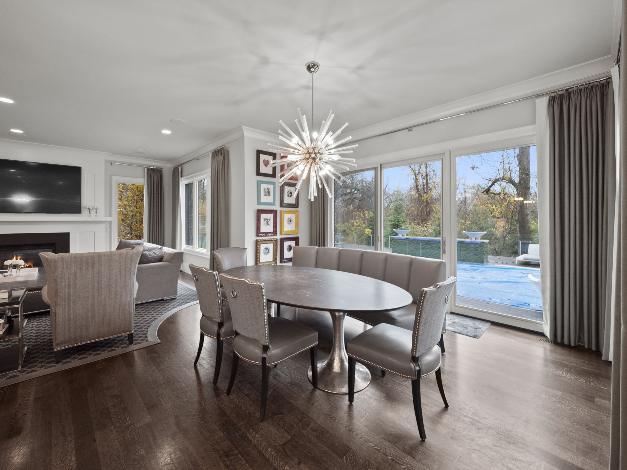 This is an interior shot of a dining room featuring a dark wood floor, an oval dining table with gray chairs and a built-in bench. A modern chandelier hangs above the table, and framed artwork adorns the wall. Large sliding glass doors offer a view of the backyard and pool area, creating a bright and inviting space.