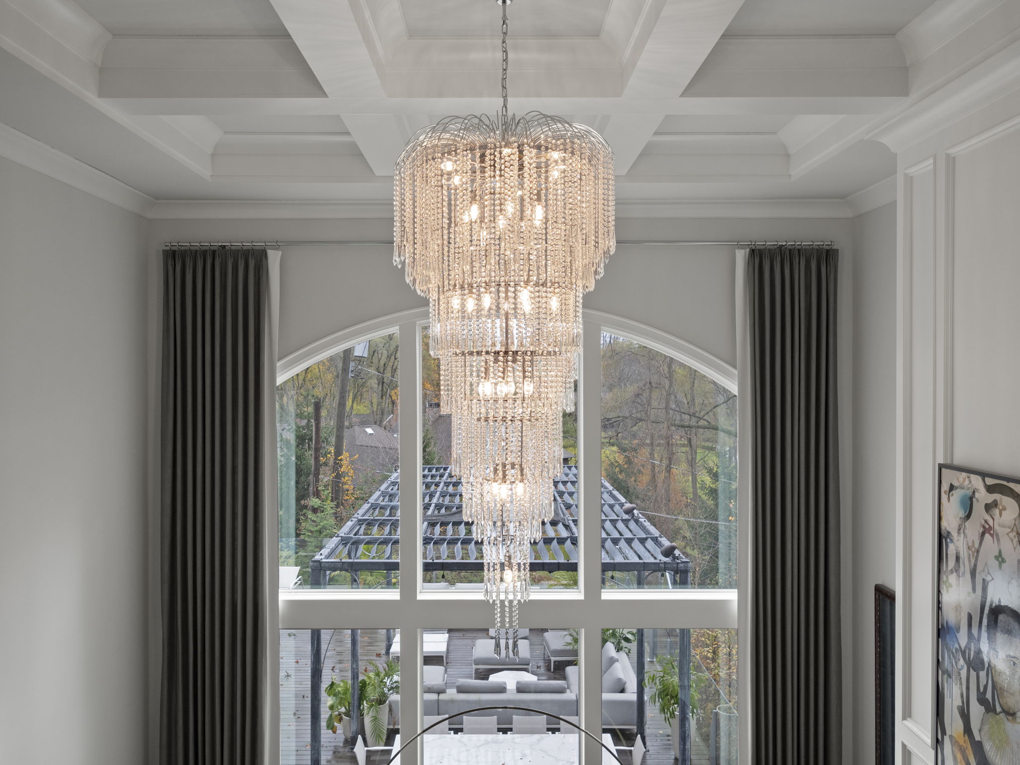 This interior shot showcases a luxurious dining room with a grand, multi-tiered crystal chandelier as the focal point. Large windows provide ample natural light and a view of the outdoor patio and garden. The room exudes elegance and sophistication, perfect for upscale dining and entertaining.