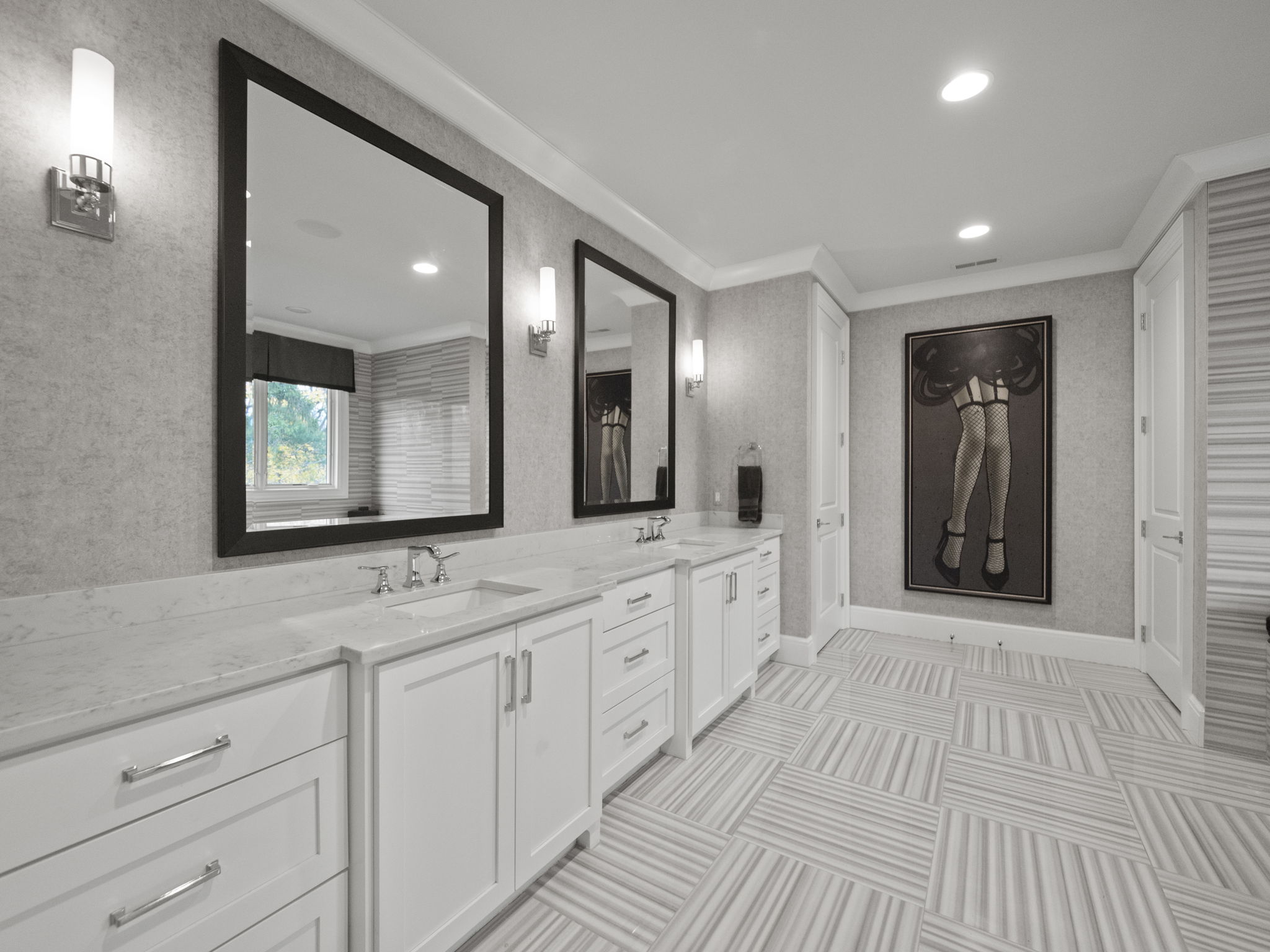 This is a well-lit primary bathroom featuring a double vanity with white cabinetry and a marble countertop. Two large mirrors with dark frames hang above the sinks, and sconces provide additional lighting. The floor is tiled in a striped pattern, and a framed art piece adds a touch of elegance to the space.