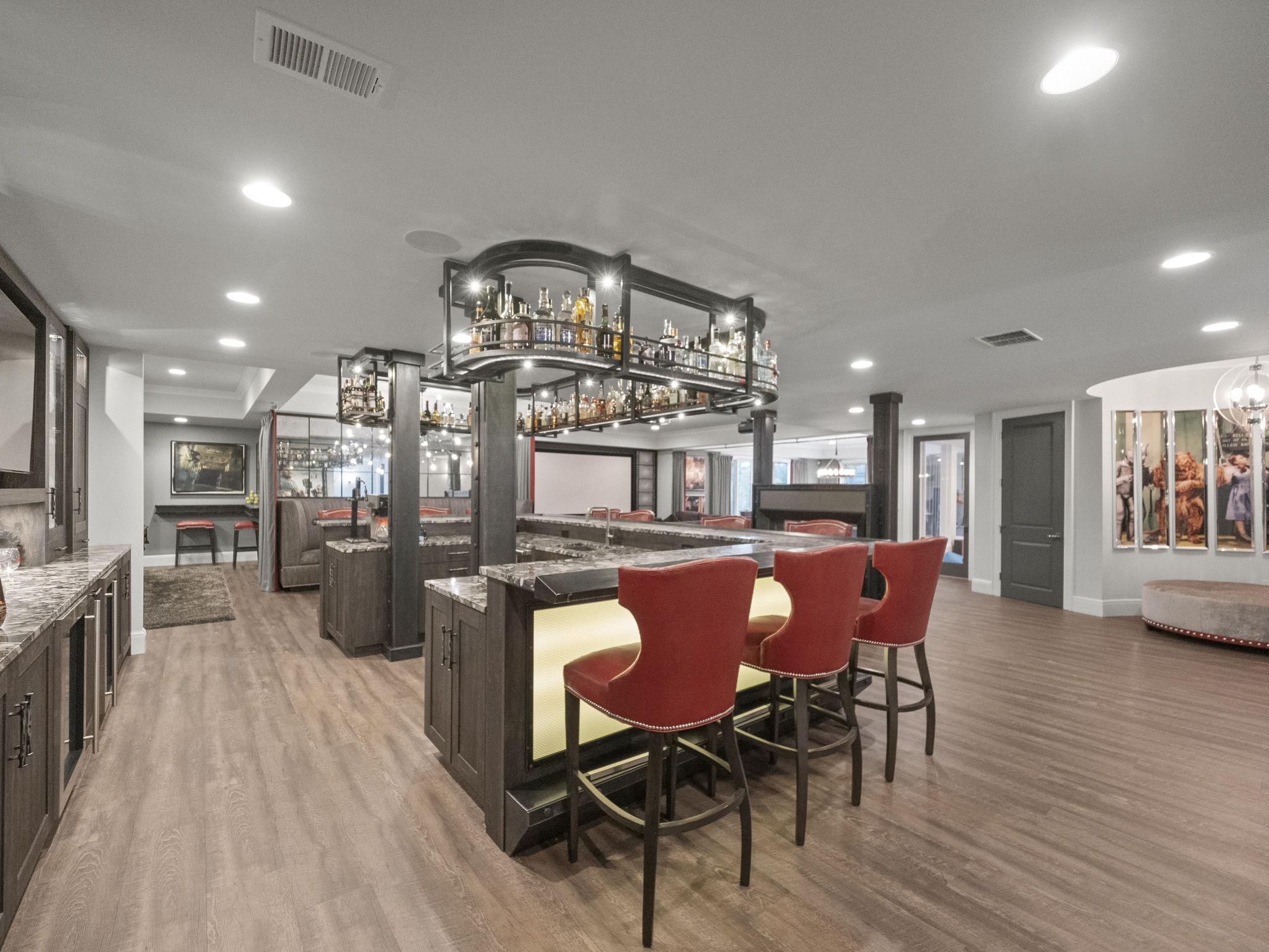 This is a spacious basement featuring a stylish bar area with red bar stools and a well-stocked liquor display. The flooring is wood-like, and the lighting is bright and modern. The overall impression is that of a luxurious and inviting entertainment space.