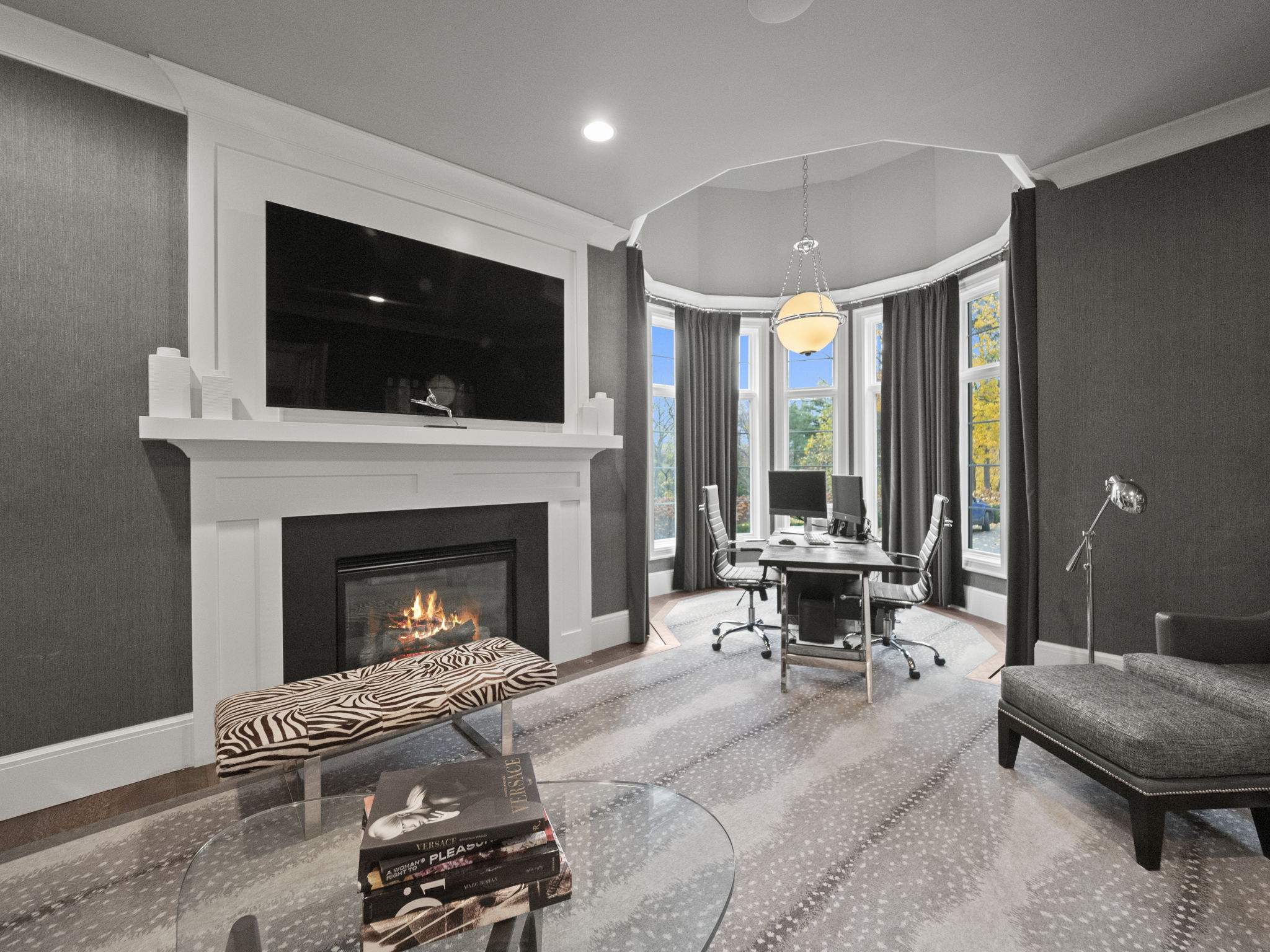 This interior shot showcases a luxurious home office featuring a fireplace with a large television mounted above. The room is decorated in a sophisticated gray palette with a patterned rug and a unique glass coffee table. A separate area with a desk and chairs is situated in a bay window, creating a bright and functional workspace.