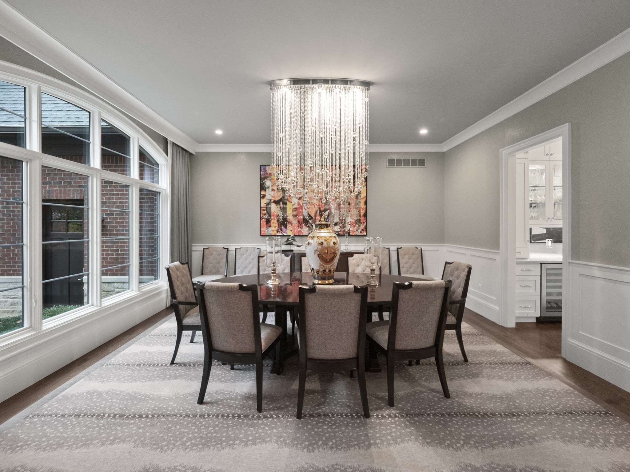 This is an interior shot of a formal dining room. The room features a large, dark wood dining table surrounded by upholstered chairs, a striking crystal chandelier, and an abstract painting on the wall. A large window provides natural light, and the room is decorated in neutral tones with elegant wainscoting, creating a sophisticated and inviting atmosphere.