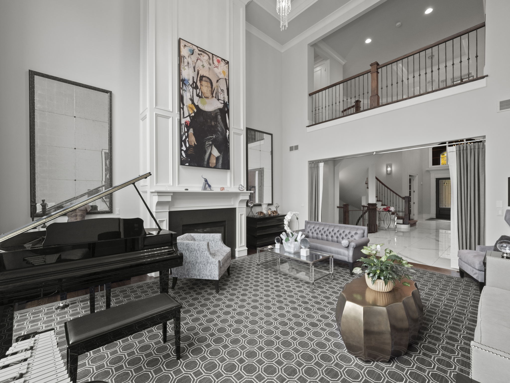 This is a grand living room featuring a black grand piano, a fireplace with a large painting above it, and elegant seating arrangements. The room boasts high ceilings and a balcony overlooking the space, creating an open and luxurious atmosphere. The flooring is patterned, and the overall style is sophisticated and modern.