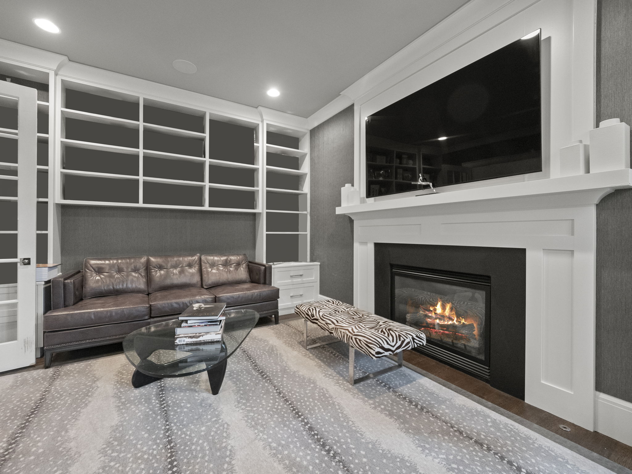 This living room features a brown leather sofa, a glass coffee table, and a zebra-striped bench, all set on a patterned rug. A fireplace with a large television above it is the focal point, flanked by built-in bookshelves. The room has a sophisticated and cozy atmosphere.