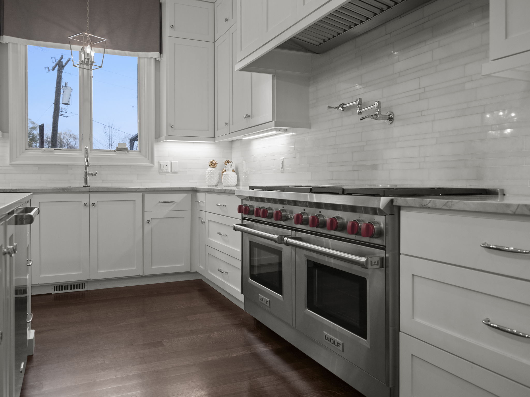 This is a well-lit kitchen featuring white cabinets, stainless steel appliances, and dark hardwood floors. A large Wolf range with red knobs is a focal point, complemented by a pot filler faucet above. The kitchen has a clean and modern aesthetic, with a window providing natural light.