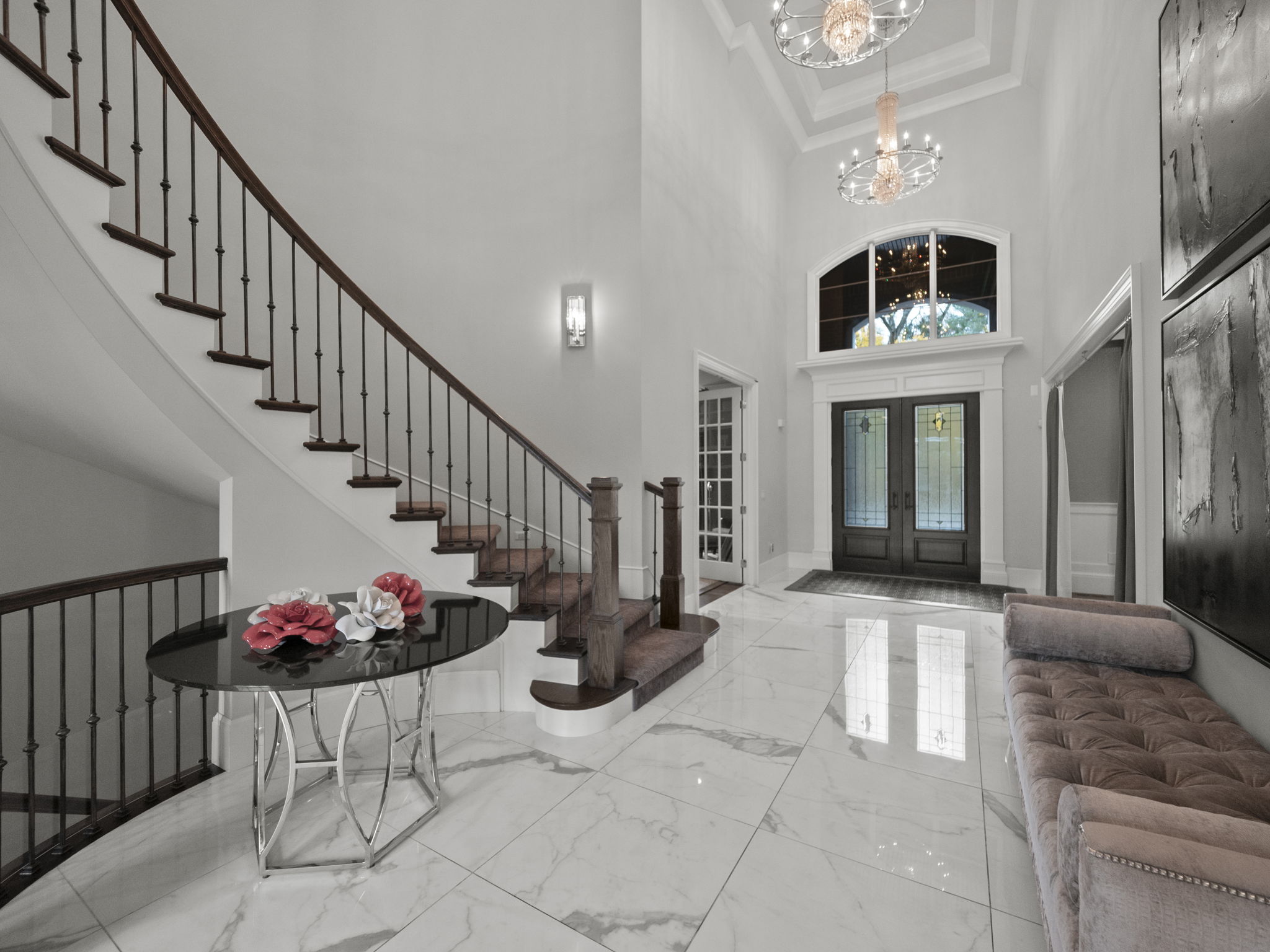 This grand foyer features a sweeping staircase with dark wood and wrought iron railings, leading to an upper level. The entrance is illuminated by elegant chandeliers and wall sconces, highlighting the marble flooring and a tufted bench. The space exudes luxury and sophistication, creating a memorable first impression.