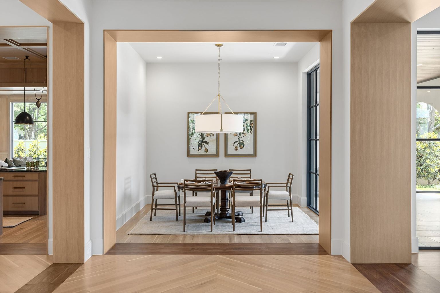 This elegant dining room is framed by a wide, light wood-trimmed archway, creating a sophisticated transition from the foreground. The space features a round dark wood table surrounded by six upholstered chairs, centered beneath a modern drum pendant light. Two framed botanical prints hang on the back wall, while large, dark-framed windows to the right provide natural light and a view of the outdoors.