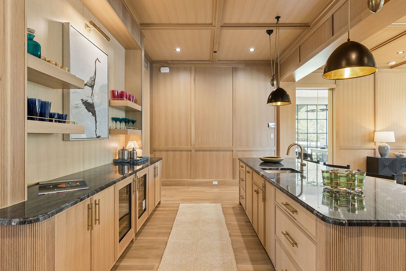 This sophisticated kitchen features light wood cabinetry with a fluted texture and dark stone countertops, creating a warm and modern aesthetic. A central island provides additional workspace and seating, complemented by hanging pendant lights and open shelving displaying glassware. The space is finished with a neutral runner rug and recessed lighting, offering a clean, high-end look suitable for entertaining.