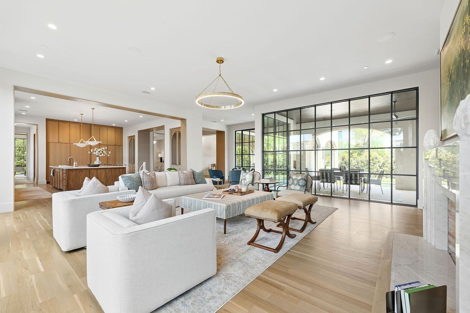 This spacious, open-concept living room features light wood flooring and a neutral color palette, creating an airy and sophisticated atmosphere. A large, grid-patterned glass wall provides a seamless transition to the outdoor patio, while a central seating area is anchored by a patterned ottoman and plush white sofas. The room flows directly into a well-appointed kitchen, highlighting a cohesive design that is ideal for modern entertaining.