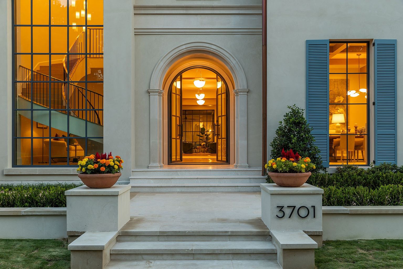 This elegant front entryway features a symmetrical facade with a grand arched doorway flanked by large, multi-pane windows. The stone steps lead up to the entrance, which is accented by two matching planters filled with vibrant flowers atop stone pedestals, one of which displays the house number 3701. The warm interior lighting creates an inviting glow, highlighting the sophisticated architectural details and the clean, light-toned exterior walls.