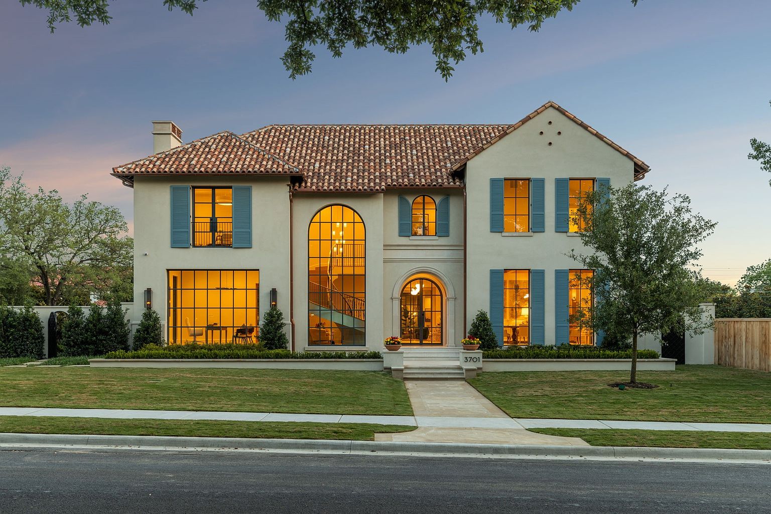 This elegant two-story Mediterranean-style home features a light-colored stucco facade, a classic terracotta tile roof, and distinctive blue shutters. The front elevation is defined by a large, arched window section that showcases an interior staircase, complemented by a symmetrical layout of tall windows and a central arched entryway. A manicured lawn and a stone walkway lead to the front door, creating a sophisticated and welcoming curb appeal.