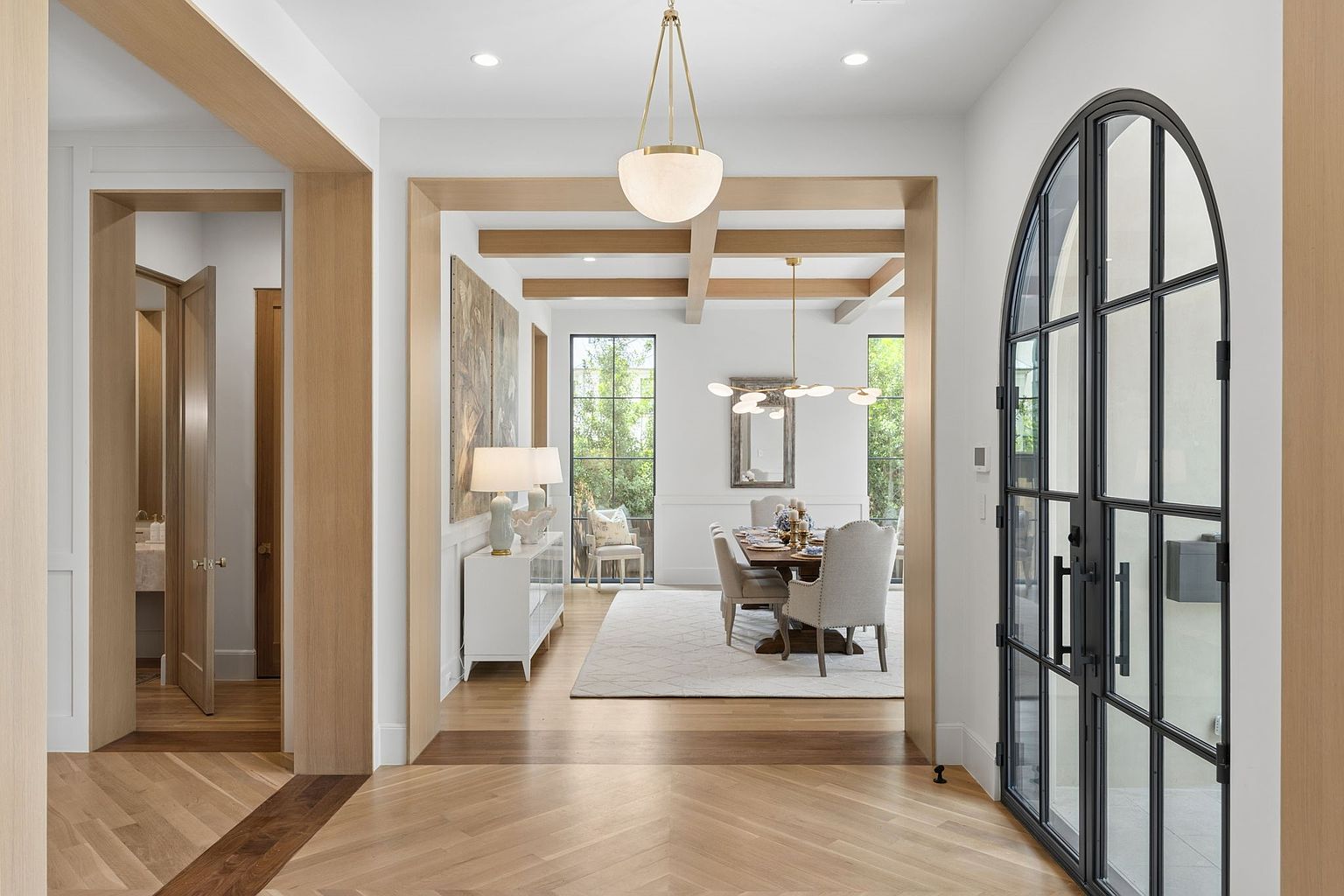 This perspective captures a bright, elegant foyer leading into a formal dining room, framed by light wood trim and architectural beams. The foreground features a herringbone-patterned wood floor and a striking arched black-framed glass door, while the dining area beyond is illuminated by a modern chandelier and large windows. The overall aesthetic is sophisticated and airy, emphasizing a seamless transition between living spaces.
