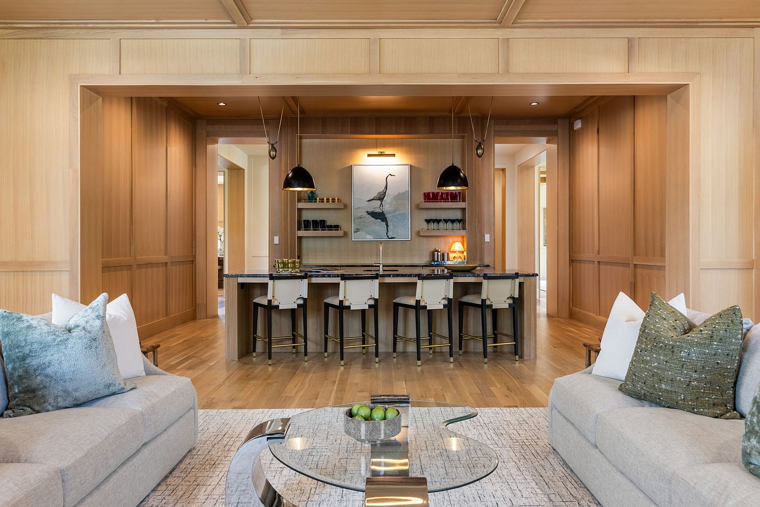 This sophisticated living space features a symmetrical layout with two plush sofas facing a central glass coffee table. Beyond the seating area, a dedicated bar nook is integrated into the light wood-paneled wall, complete with a dark stone-topped island, four modern bar stools, and decorative shelving. The warm wood tones and balanced lighting create an inviting, high-end atmosphere perfect for entertaining.