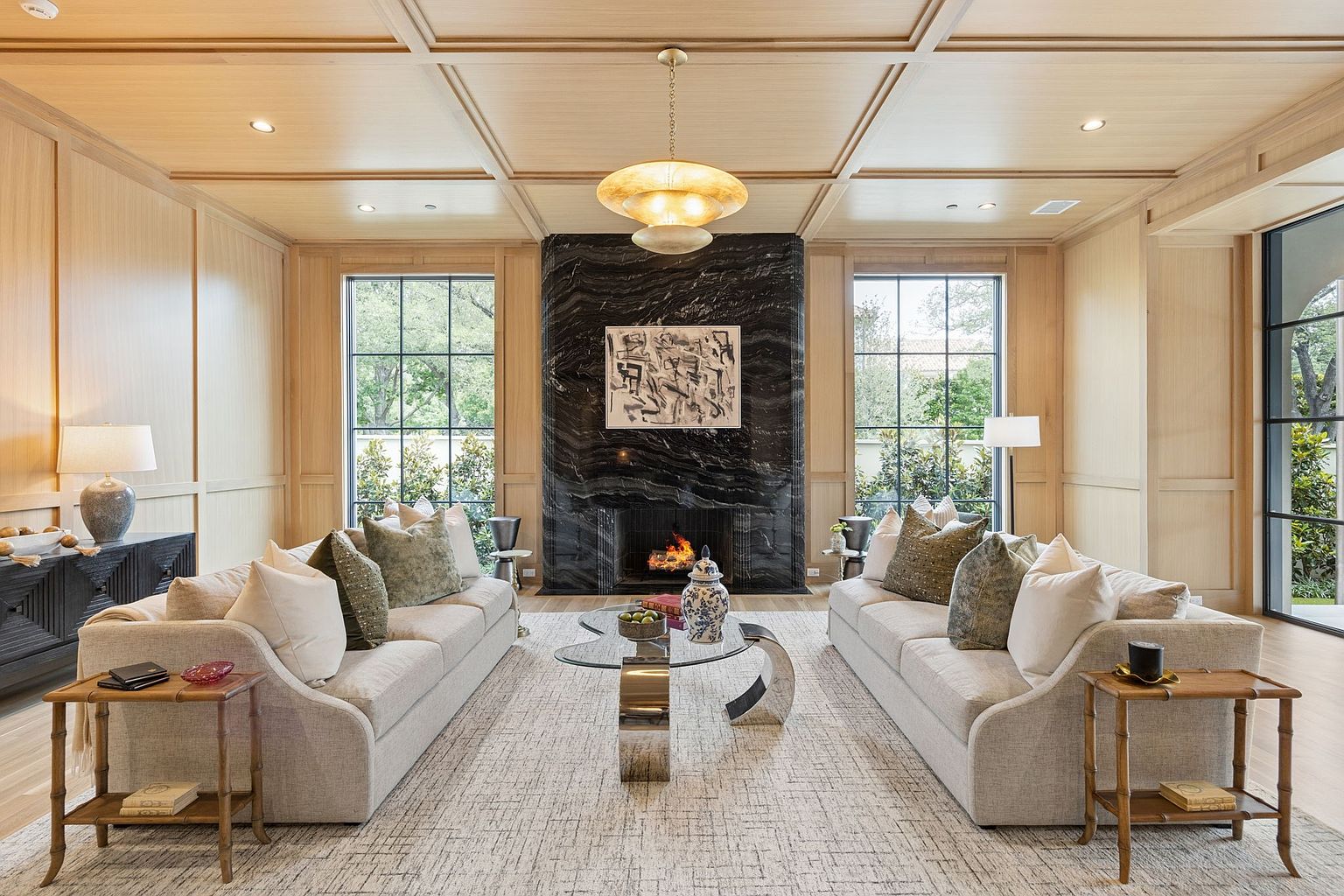 This elegant living room features a symmetrical layout centered around a striking black stone-clad chimney breast with a built-in fireplace. The space is framed by light wood paneling on the walls and ceiling, complemented by two large windows that provide ample natural light. Two matching sofas face each other across a glass-topped coffee table, creating a sophisticated and balanced atmosphere.