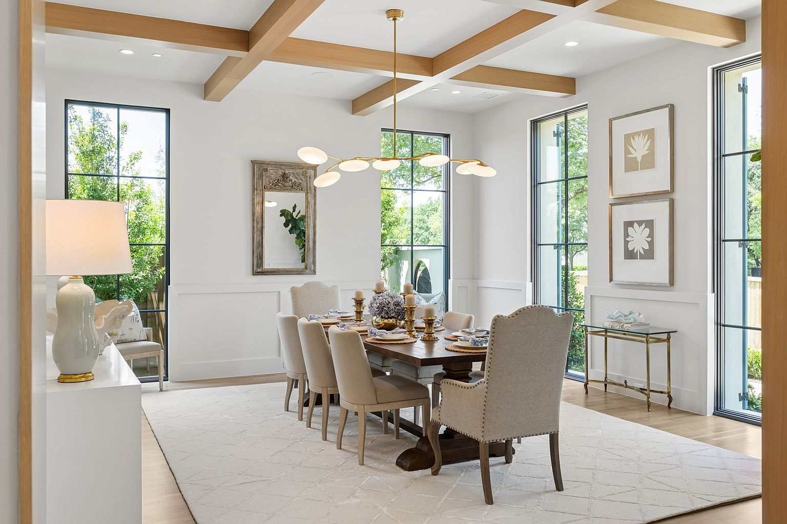 This elegant dining room features a large wooden table surrounded by upholstered chairs, set beneath a modern, multi-armed brass chandelier. The space is defined by light wood ceiling beams, tall windows with black frames that allow for abundant natural light, and a neutral-toned area rug. A decorative mirror and framed botanical art add a sophisticated touch to the bright, airy atmosphere.