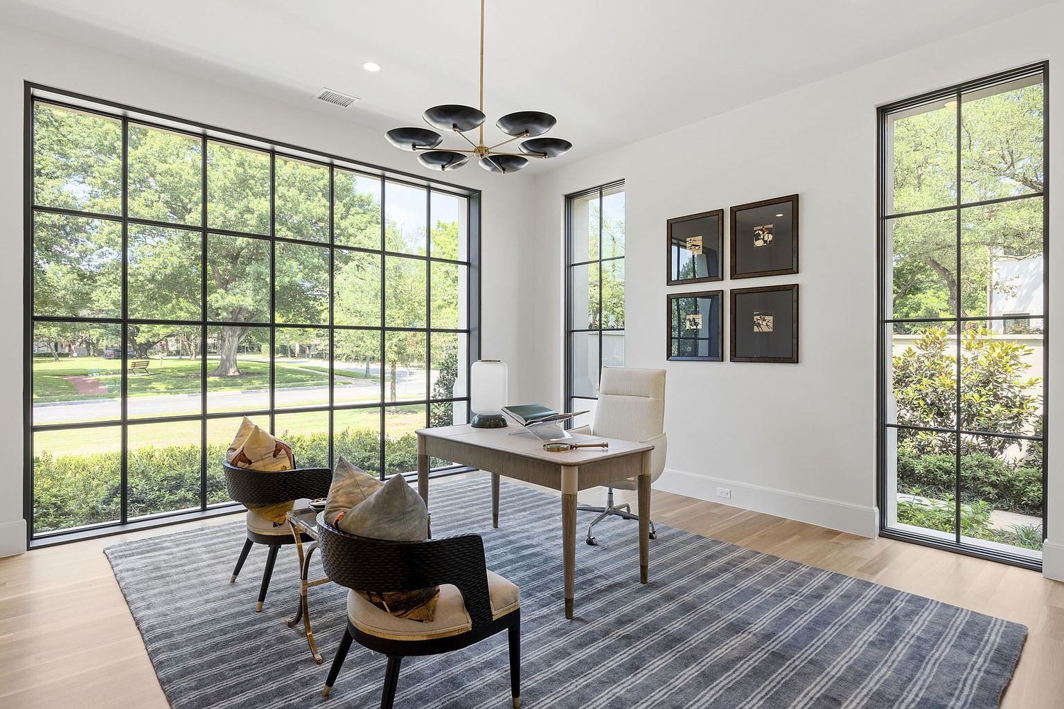 This bright and airy home office features expansive, grid-patterned windows that provide a panoramic view of the surrounding greenery. The room is anchored by a sleek, light-toned desk and a modern office chair, complemented by two dark-framed accent chairs on a textured, striped area rug. A contemporary multi-arm chandelier hangs from the ceiling, while a gallery of four framed artworks adds a sophisticated touch to the minimalist white walls.