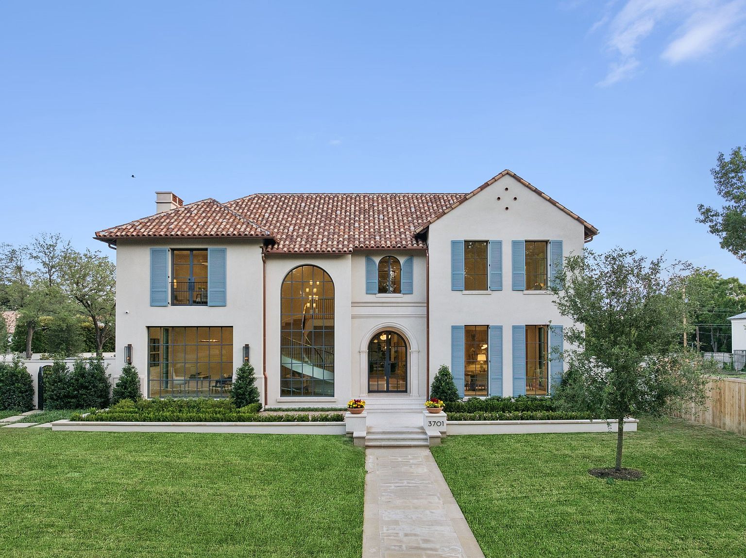 This elegant two-story residence features a Mediterranean-inspired aesthetic with a light-colored stucco facade, a terracotta tile roof, and distinctive blue shutters. The front elevation is defined by a large arched window showcasing an interior staircase, a central arched entryway, and symmetrical window placements. A manicured lawn and a stone walkway lead directly to the home, creating a welcoming and sophisticated curb appeal.
