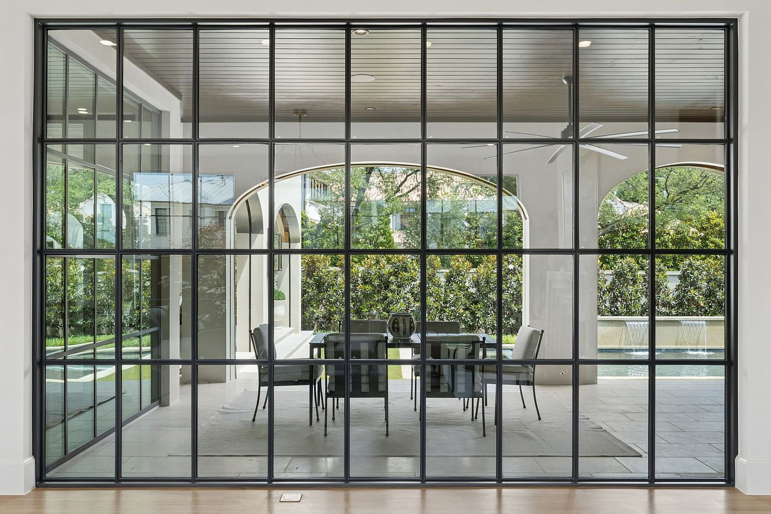 This interior view looks through a massive, grid-patterned black metal-framed glass wall into a covered outdoor dining area. The space beyond features a dining table with chairs, a ceiling fan, and an arched architectural opening that frames a view of a lush garden and a swimming pool with a water feature. The perspective emphasizes the seamless transition between the indoor living area and the sophisticated outdoor living space.
